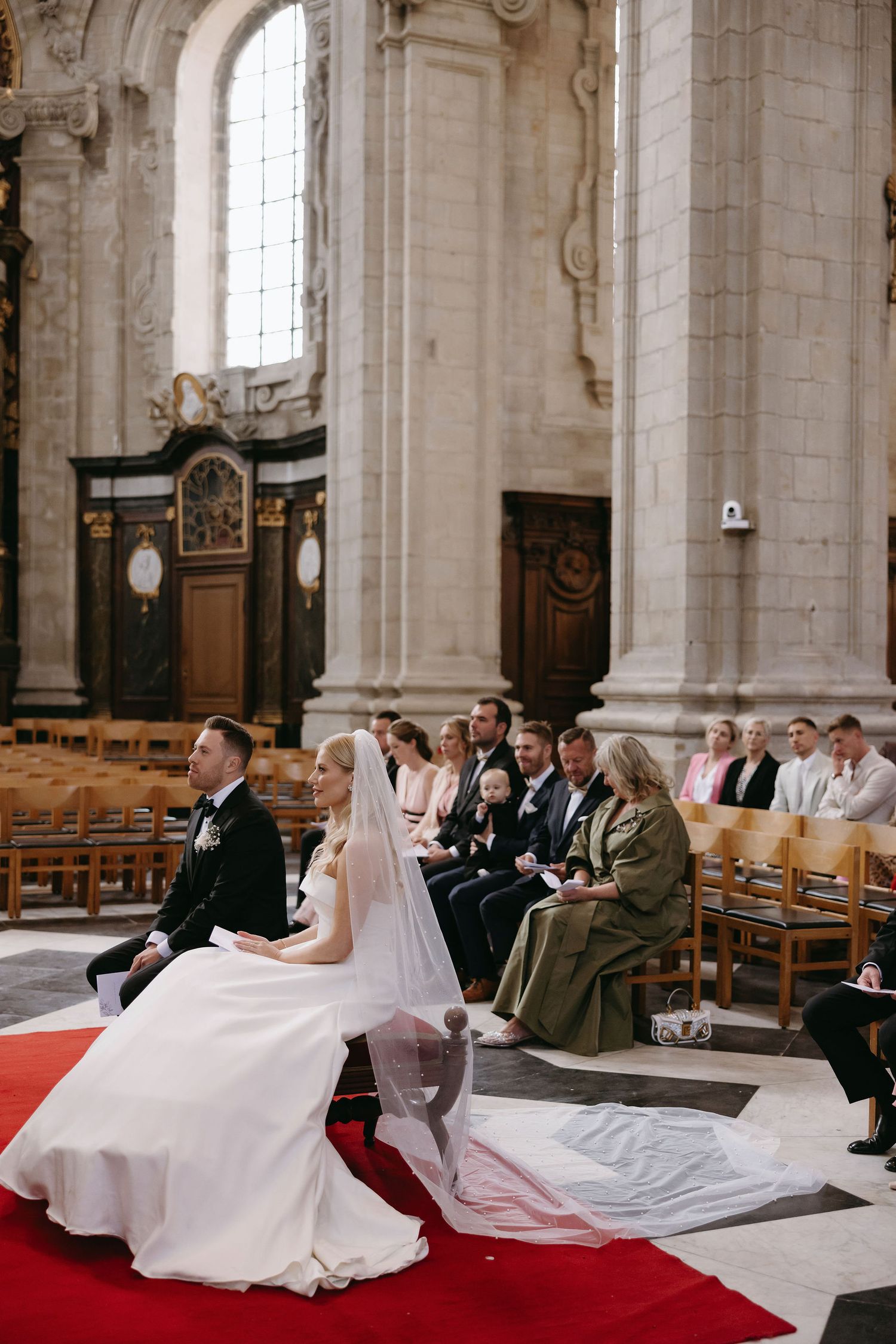 Couple kneels at altar during wedding ceremony in historic church with red carpet.