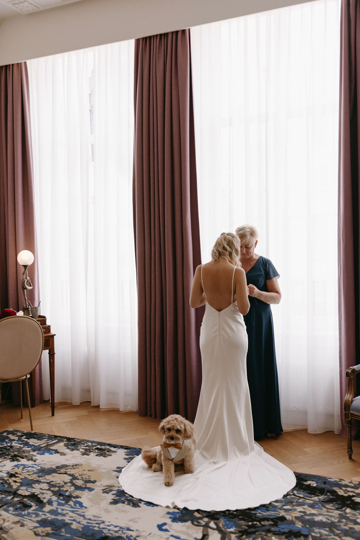 A wedding scene in a bright room with flowing curtains and a small dog sitting by a long white dress train.
