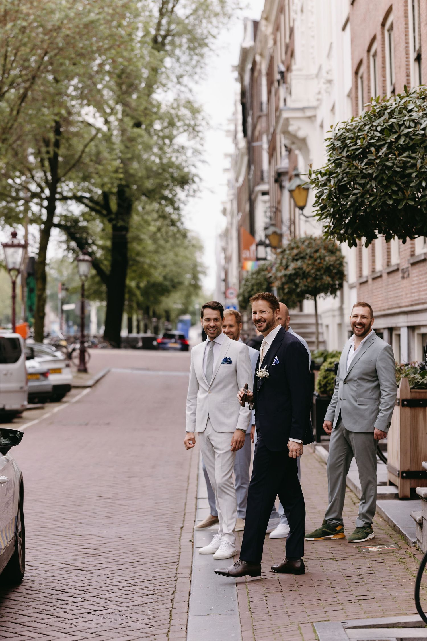 Three men in formal attire stand on a tree-lined city street with brick buildings.