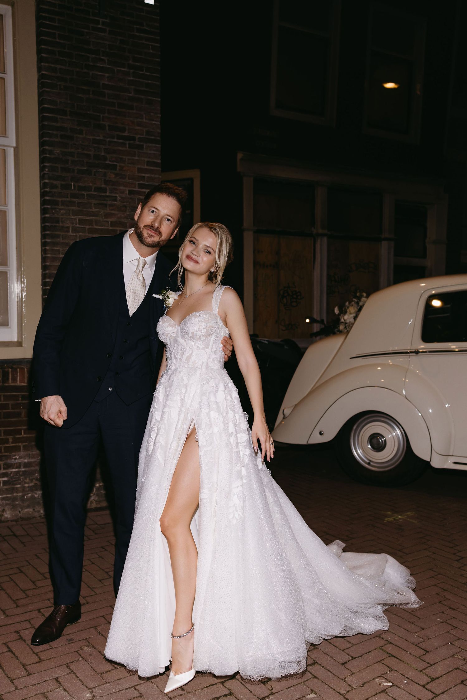 A wedding couple poses at night next to a vintage cream-colored car on brick pavement.