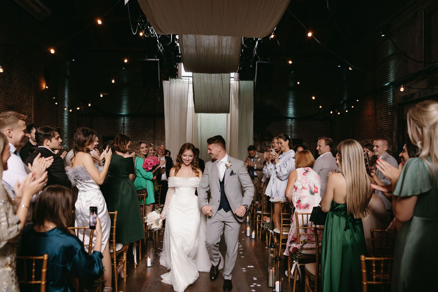 Wedding party making their grand exit through an aisle lined with guests and string lights.