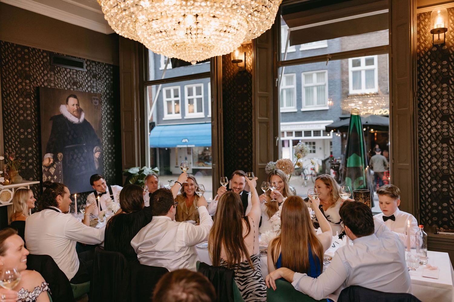 Wedding guests gathered around tables under an elegant chandelier in a restaurant setting.