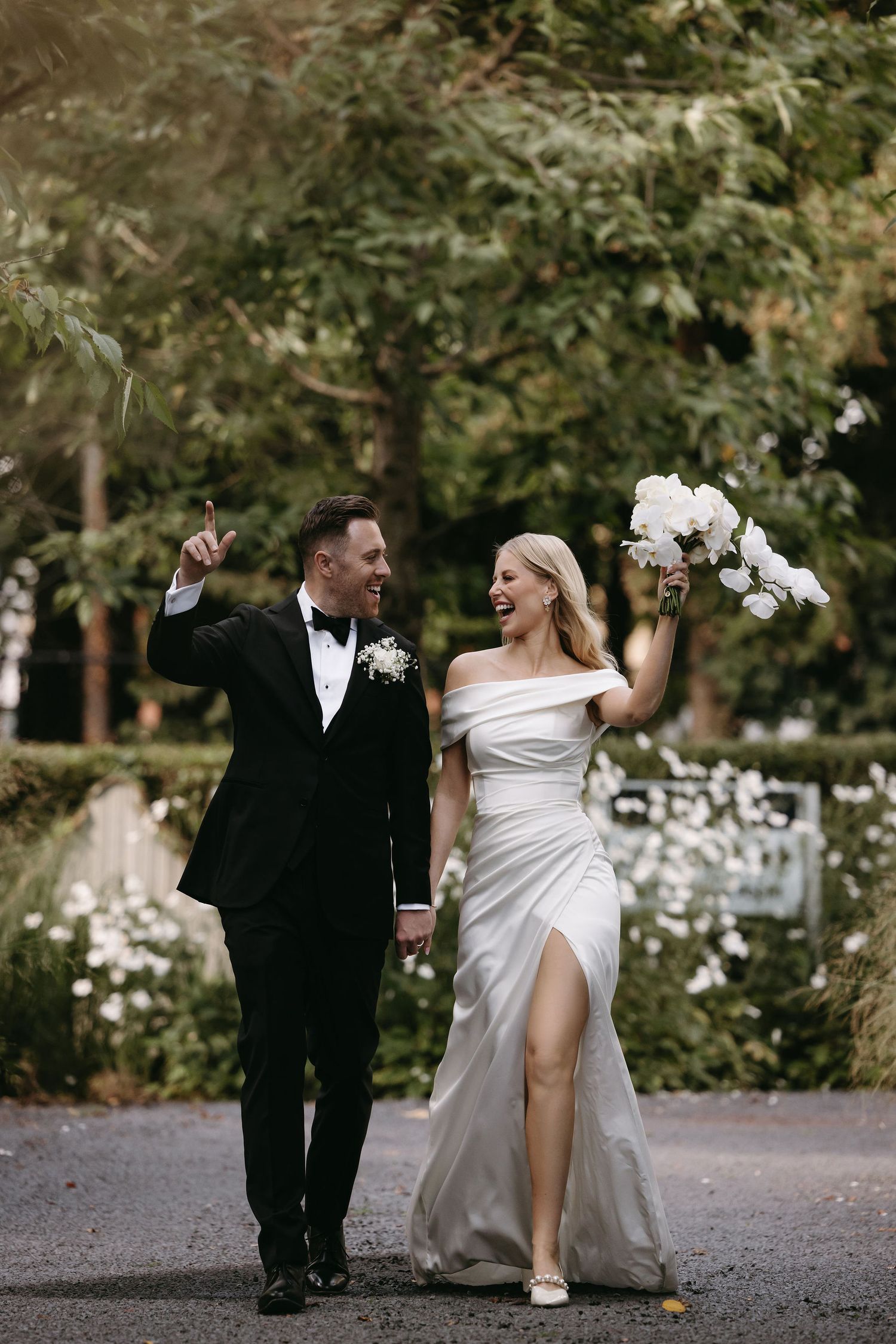 Newlyweds walking and laughing together on a garden path in formal wedding attire.