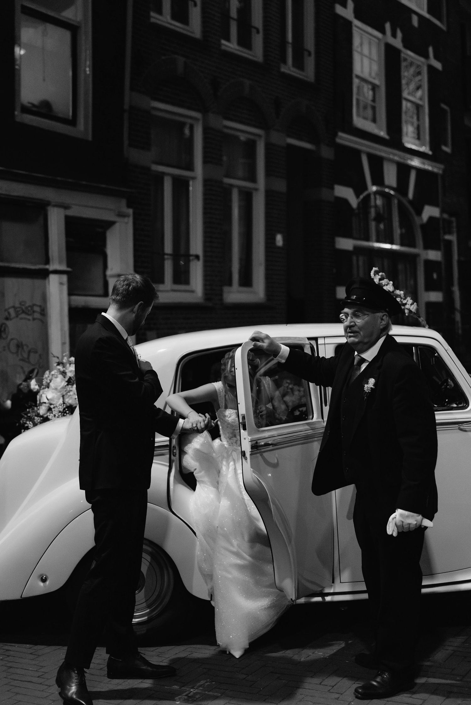 Vintage black and white photo of classic car arrival at wedding with formal attire.