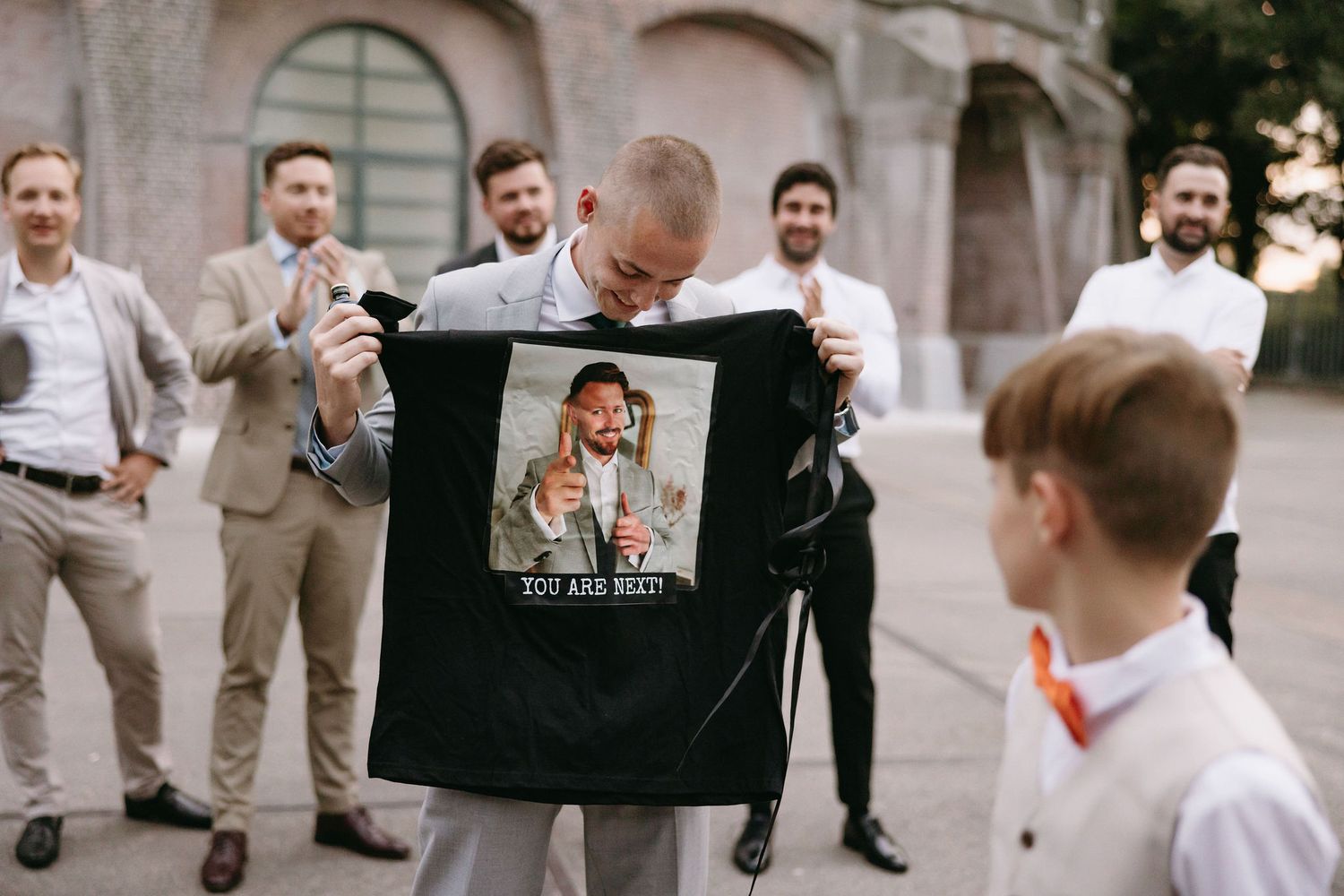 Wedding party members pose and laugh together outside a historic stone building.