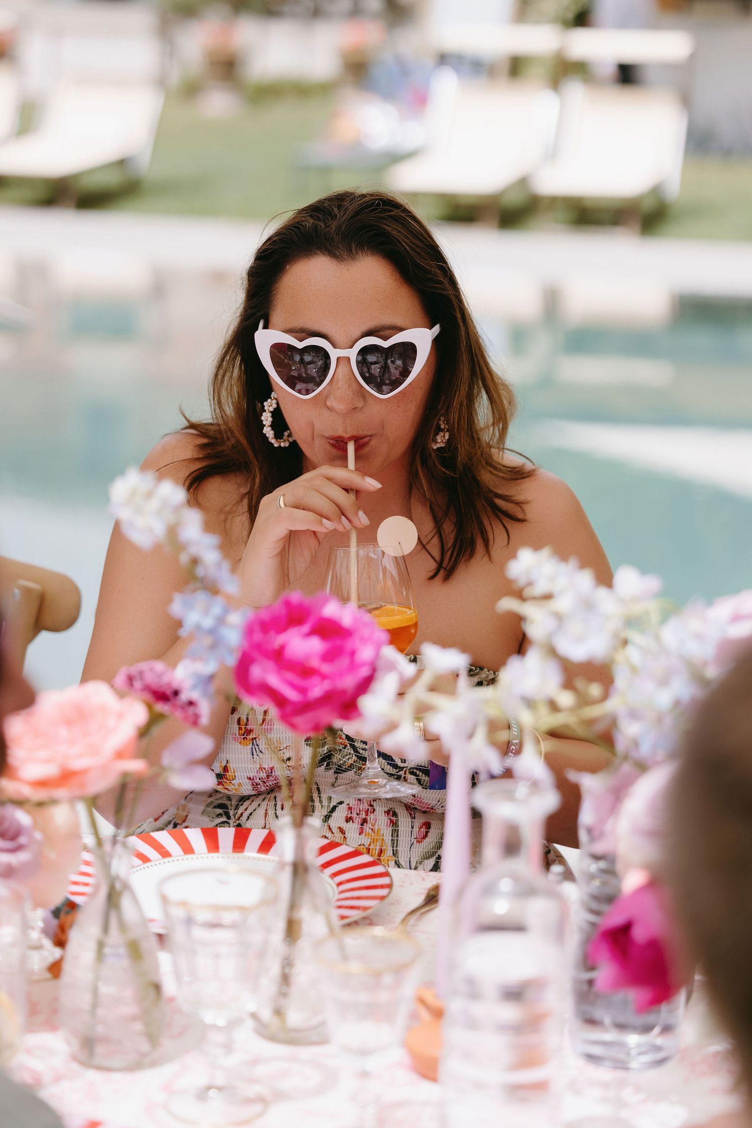 Guest in heart-shaped sunglasses sips a drink at a poolside party with pink floral decorations.