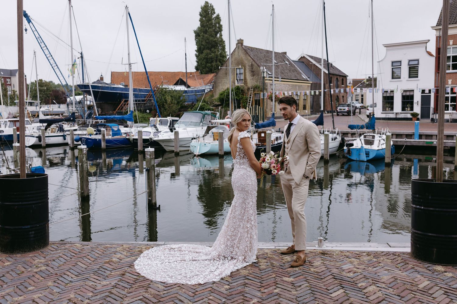 A wedding couple poses on a dock in front of boats at a marina with brick walkways and historic buildings.
