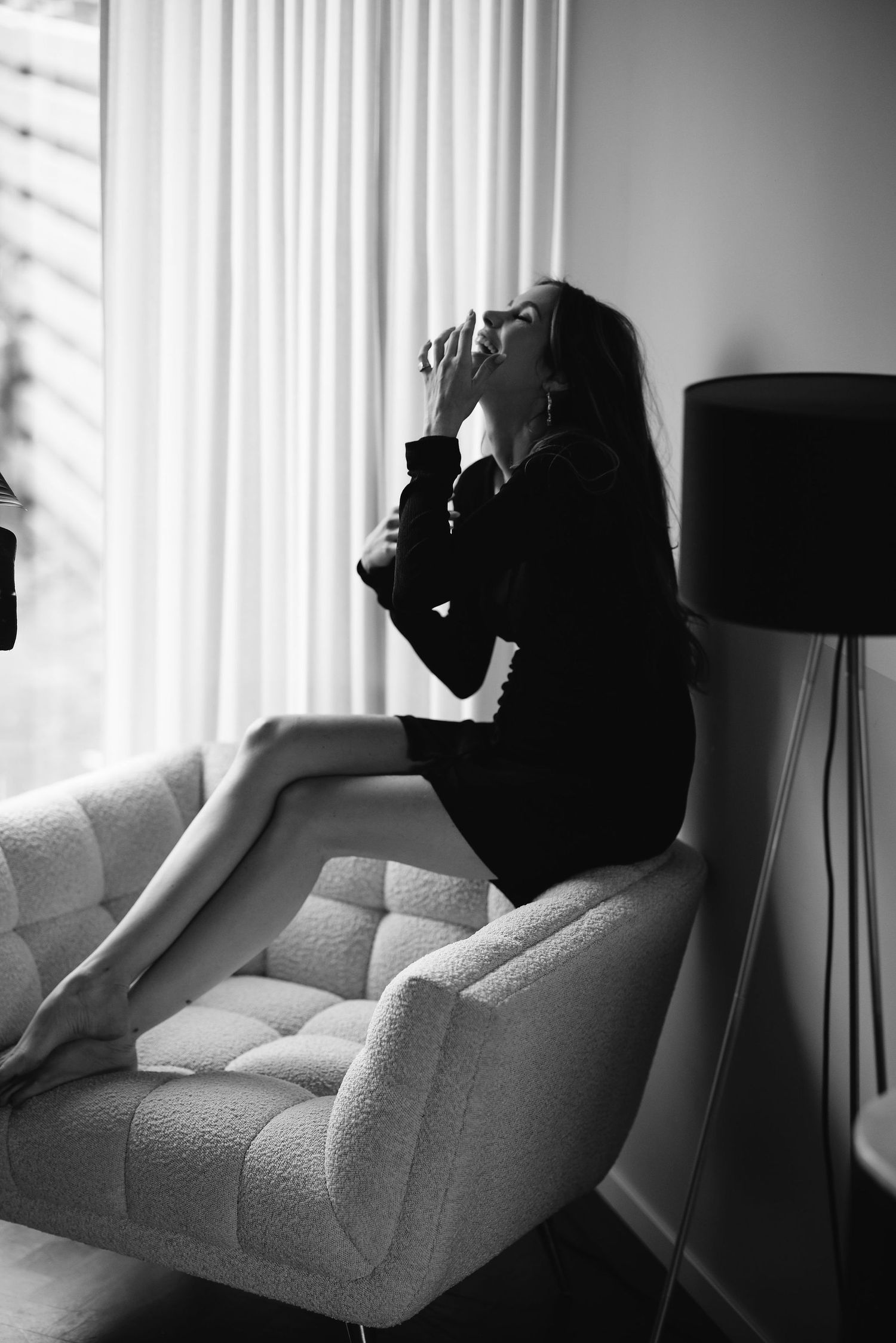 Woman in black dress reclines on gray couch near window with dramatic lighting creating moody atmosphere.