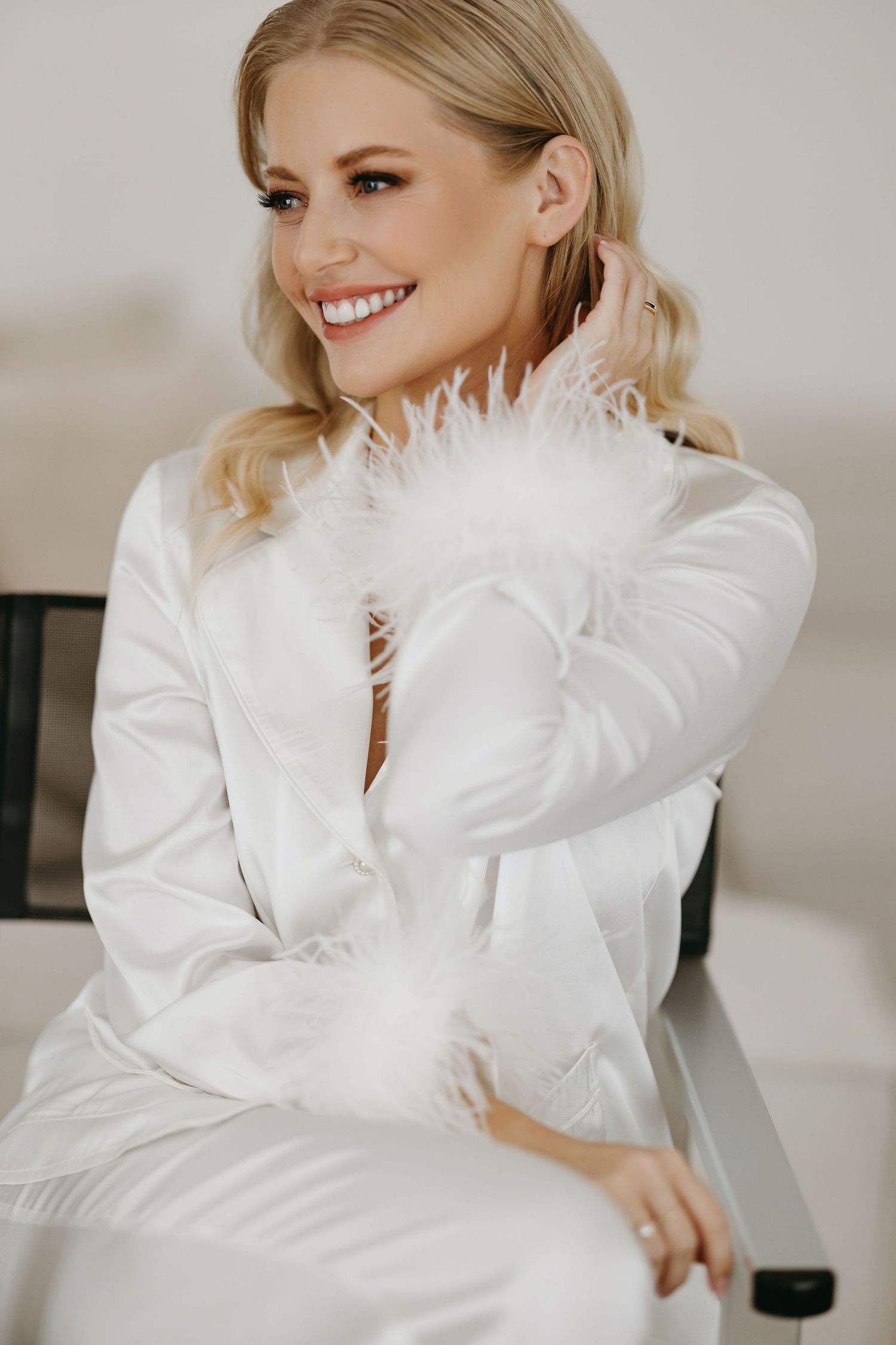 Fashion portrait of elegant model in white feather-trimmed outfit sitting indoors with bright smile.