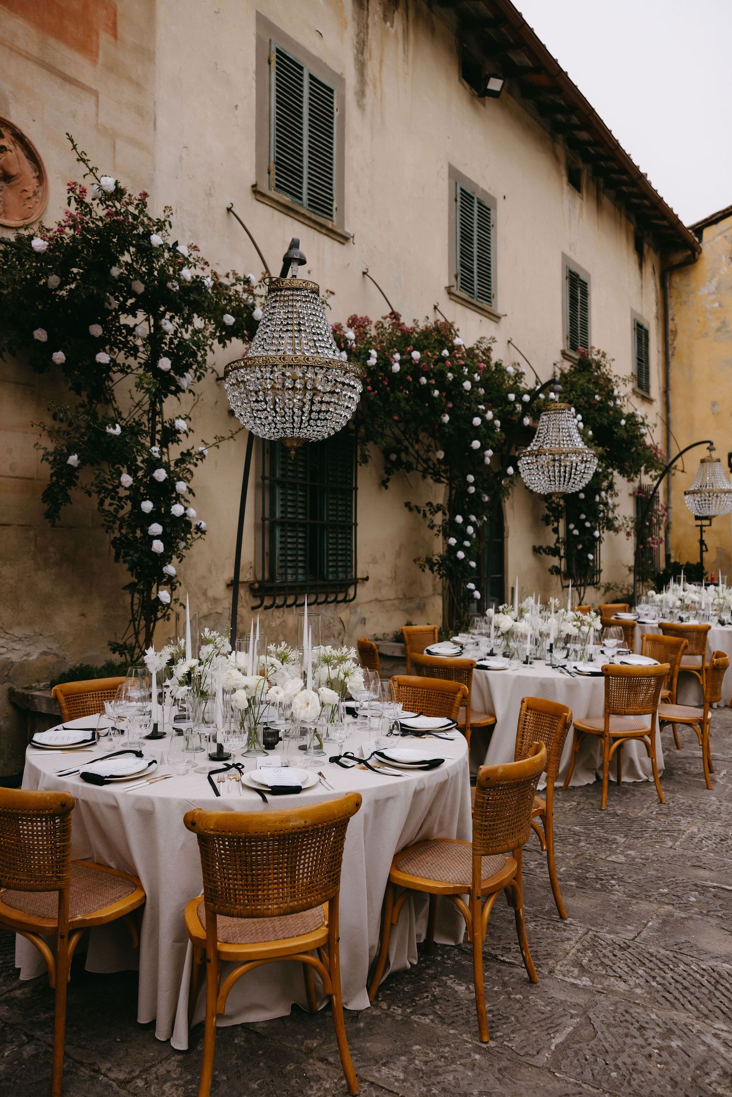 Elegant outdoor wedding reception setup with crystal chandeliers, white tablecloths and wooden chairs along a rustic wall.