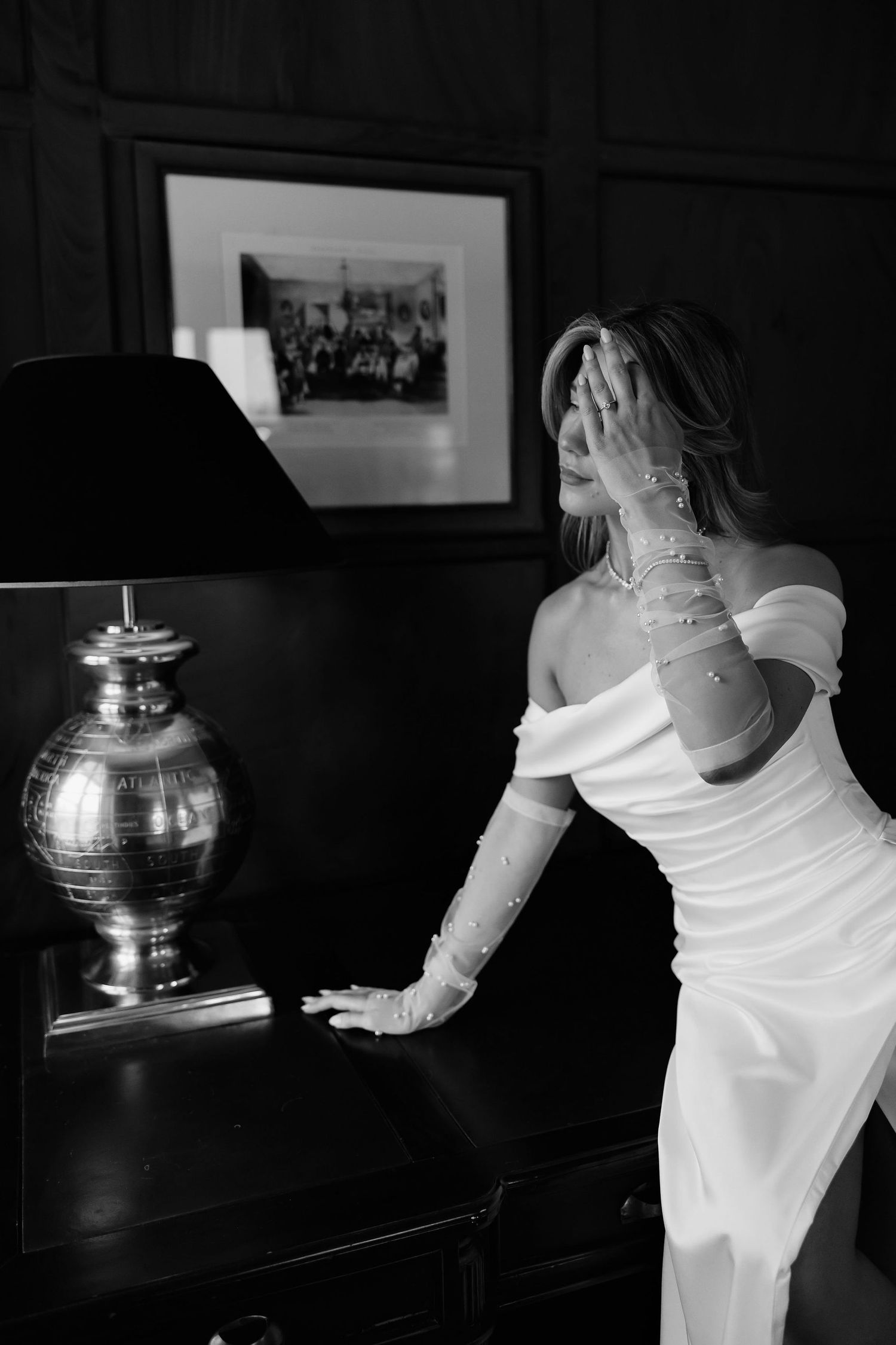 Elegant bride in off-shoulder white gown and long gloves poses dramatically next to metallic silver lamp in black and white photo.