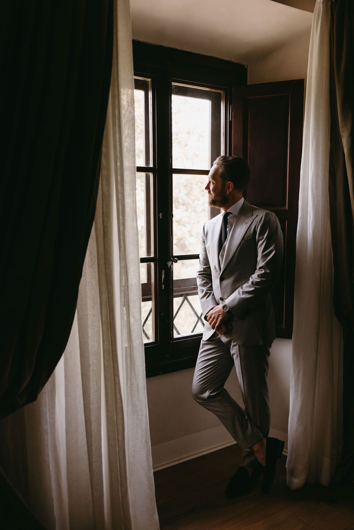 A moody portrait of someone wearing a gray suit standing by a window with white curtains in dramatic lighting.