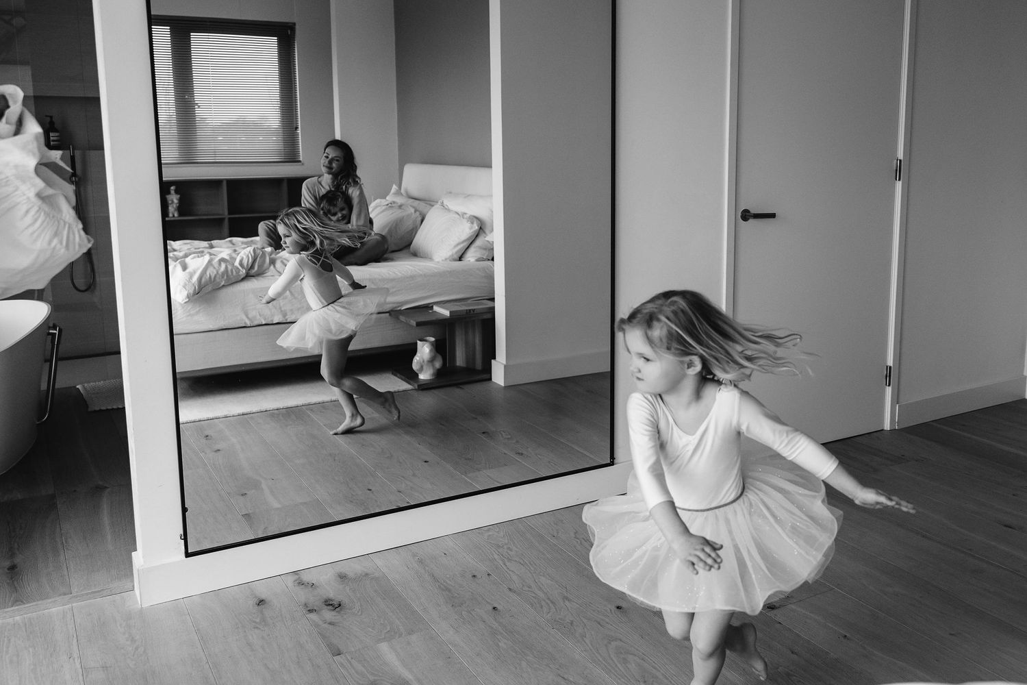 A child in white twirls in front of a mirror while parents watch from the bed.