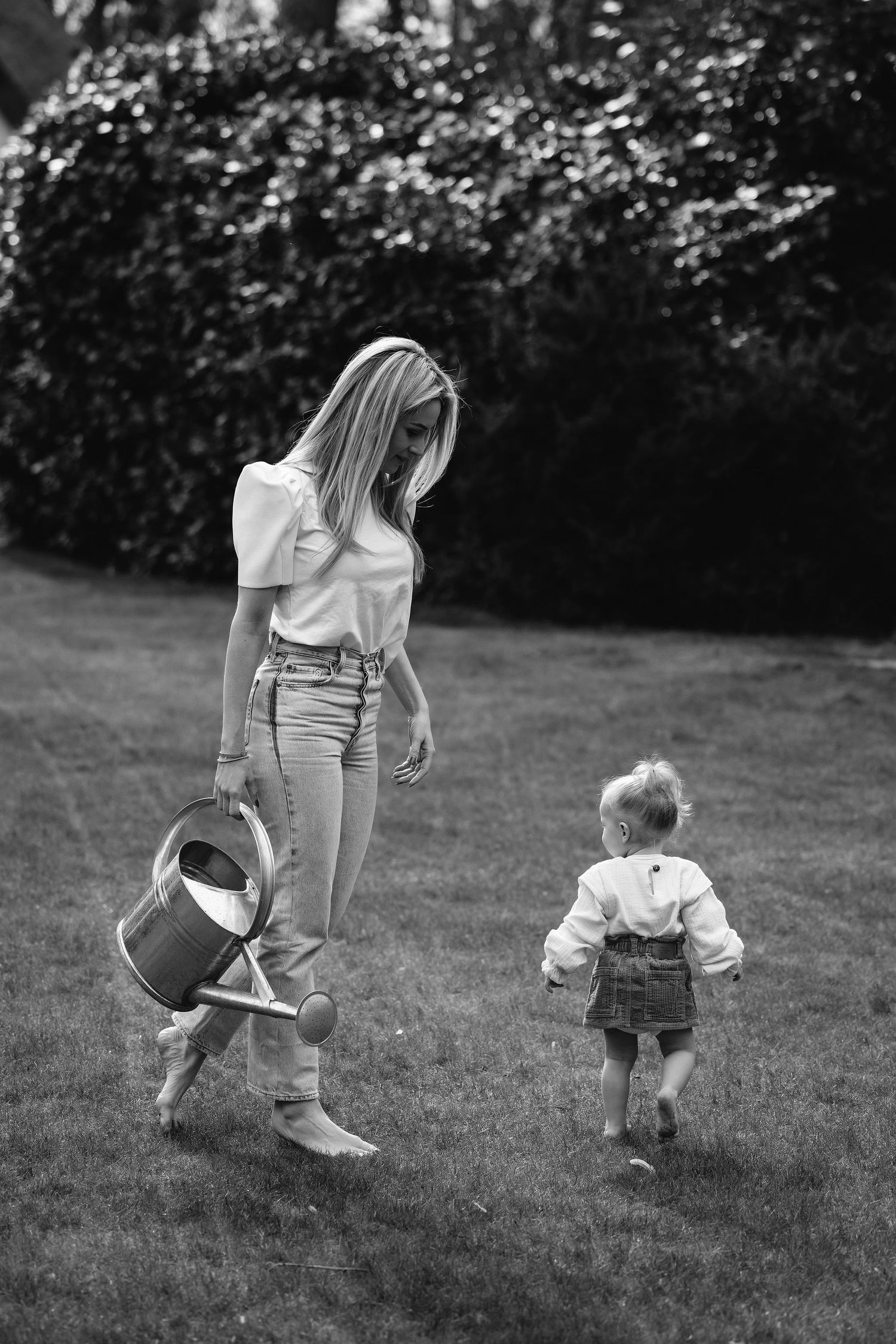 Black and white image of garden scene with watering can and toddler walking on lawn.