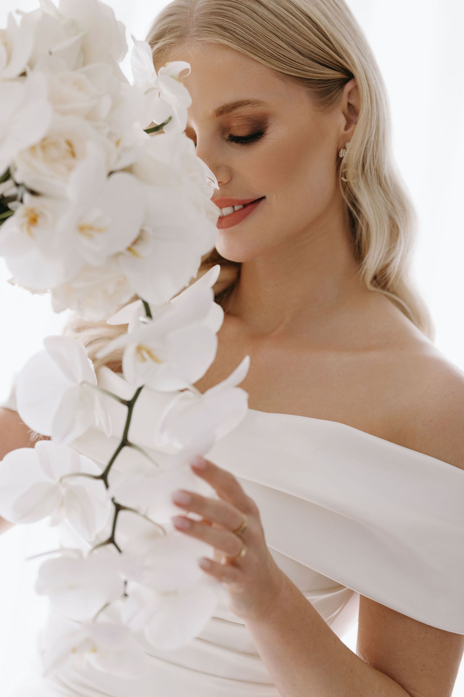 Elegant bride in off-shoulder dress holding white orchids against light background.