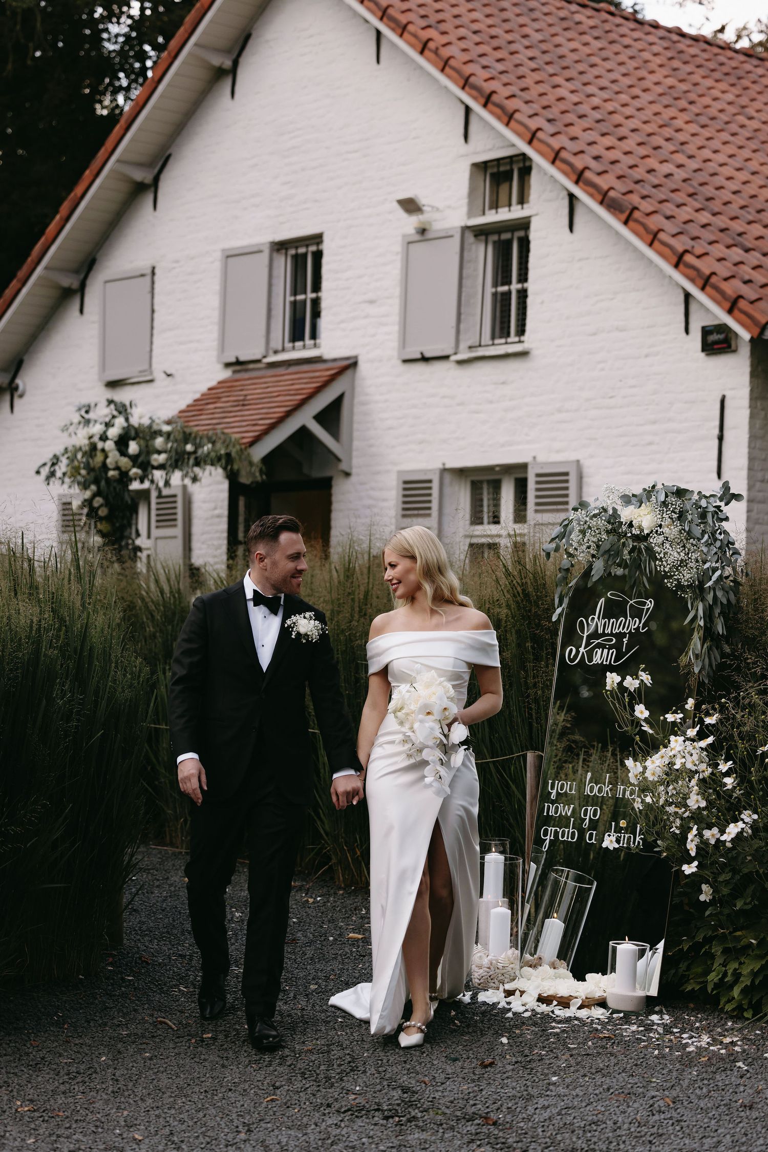 A couple walks together on a path beside a white house with terracotta roof tiles and decorative window shutters.