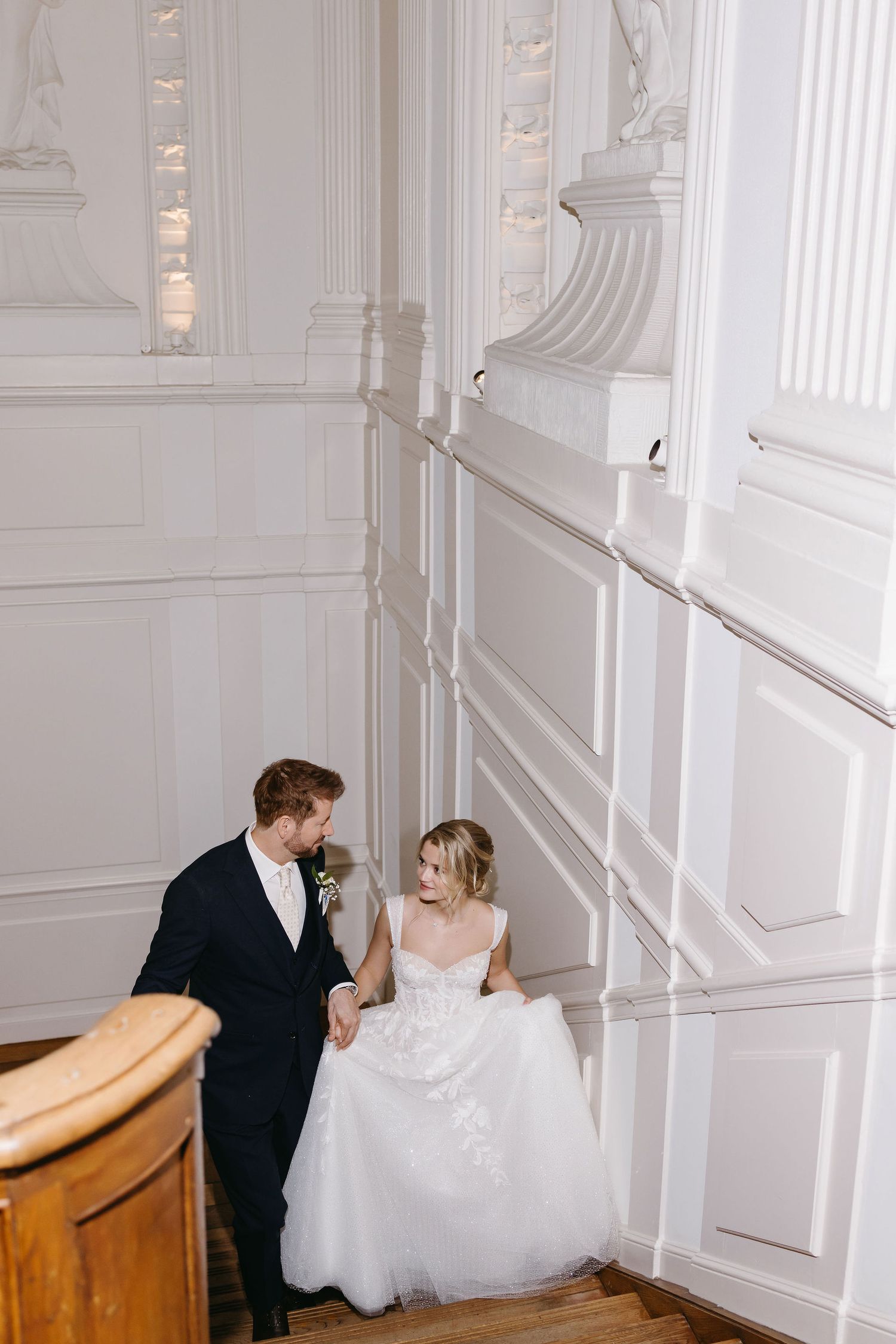 A couple descends an elegant white staircase on their wedding day surrounded by ornate architectural details.