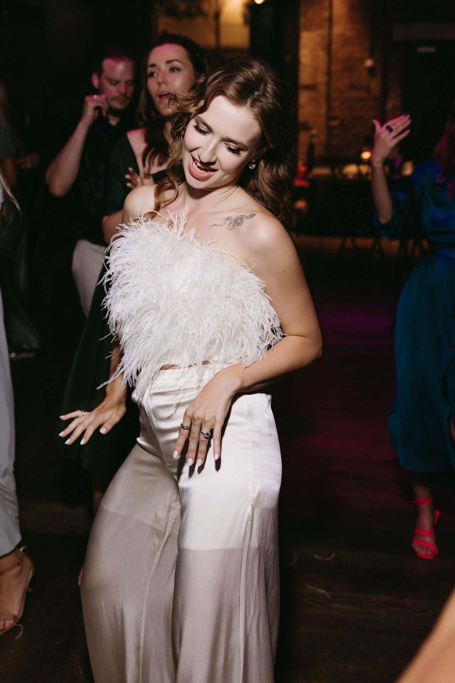 Wedding guests dancing joyfully at reception with feathered white dress detail visible.