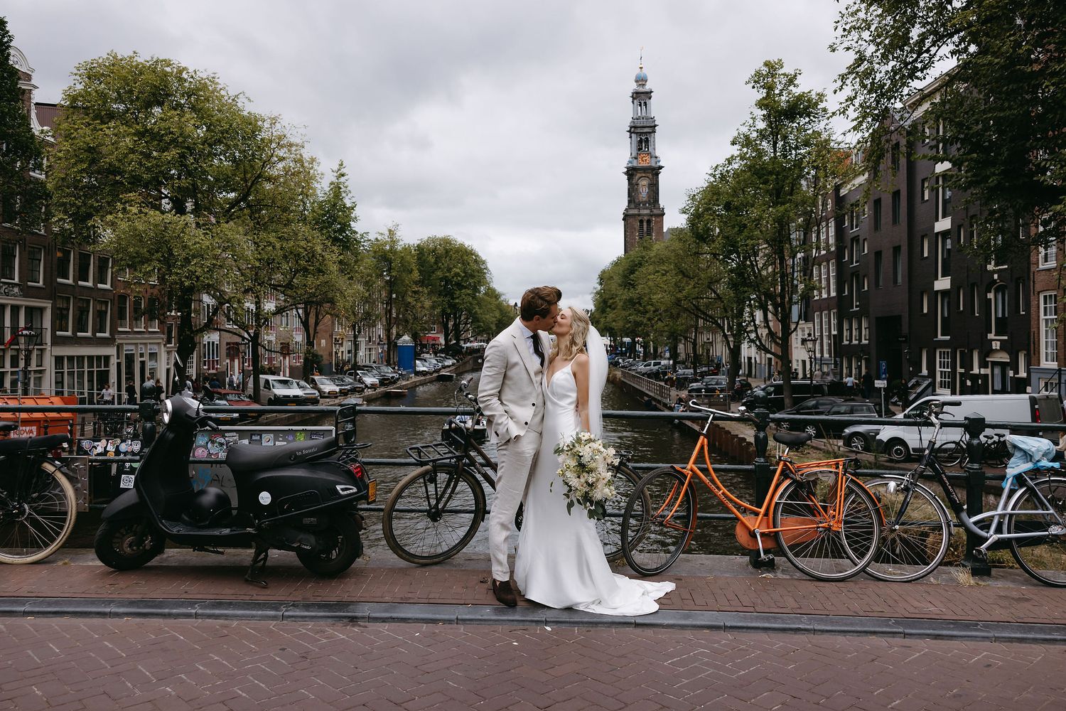 Wedding couple embracing on Amsterdam canal bridge with bicycles and classic architecture in background.