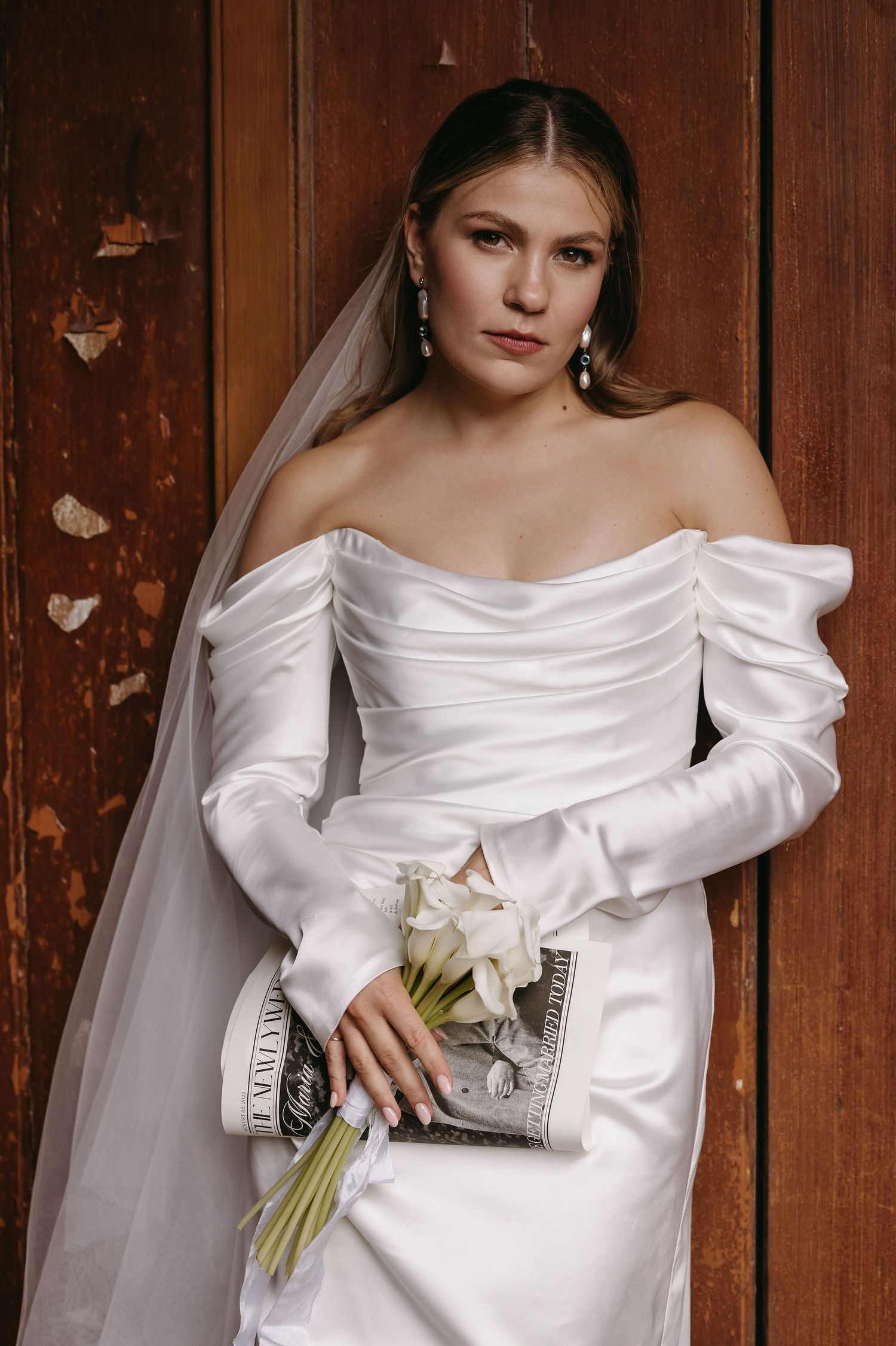 Bride in an elegant off-shoulder white wedding dress holding a bouquet against a wooden door background.