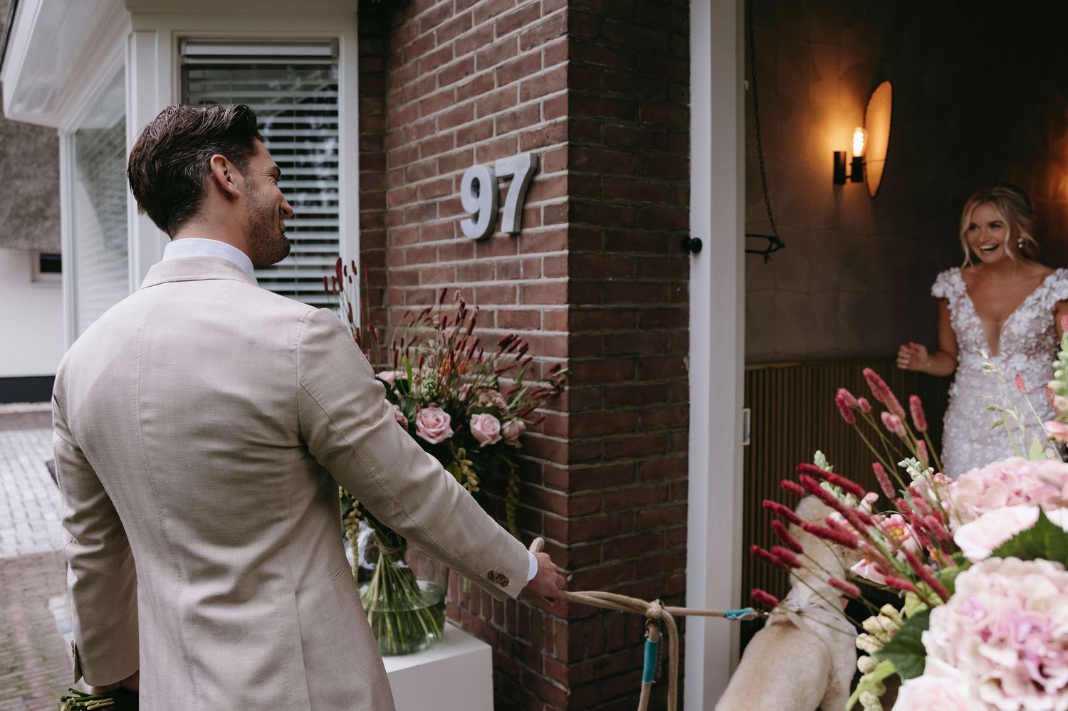 Groom with flowers stands at doorway of house number 97 during wedding day moment.