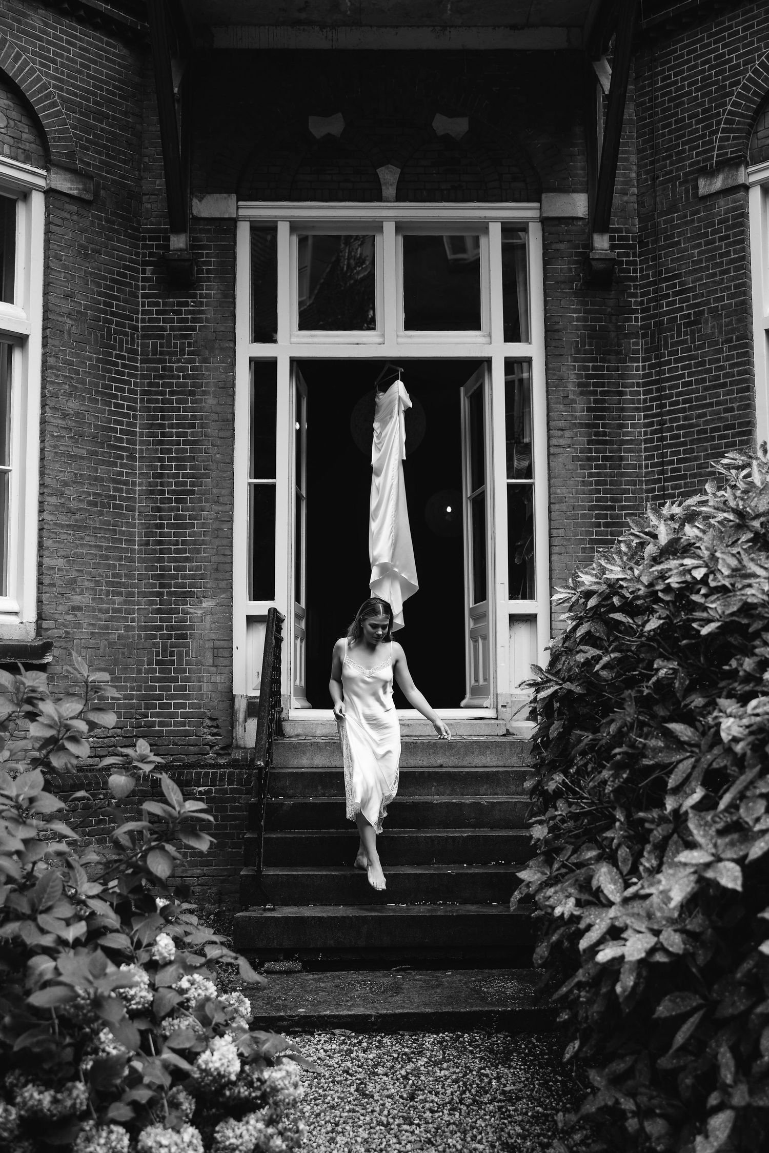 A figure in a white dress descends stone steps at the entrance of a brick building with foliage on either side.