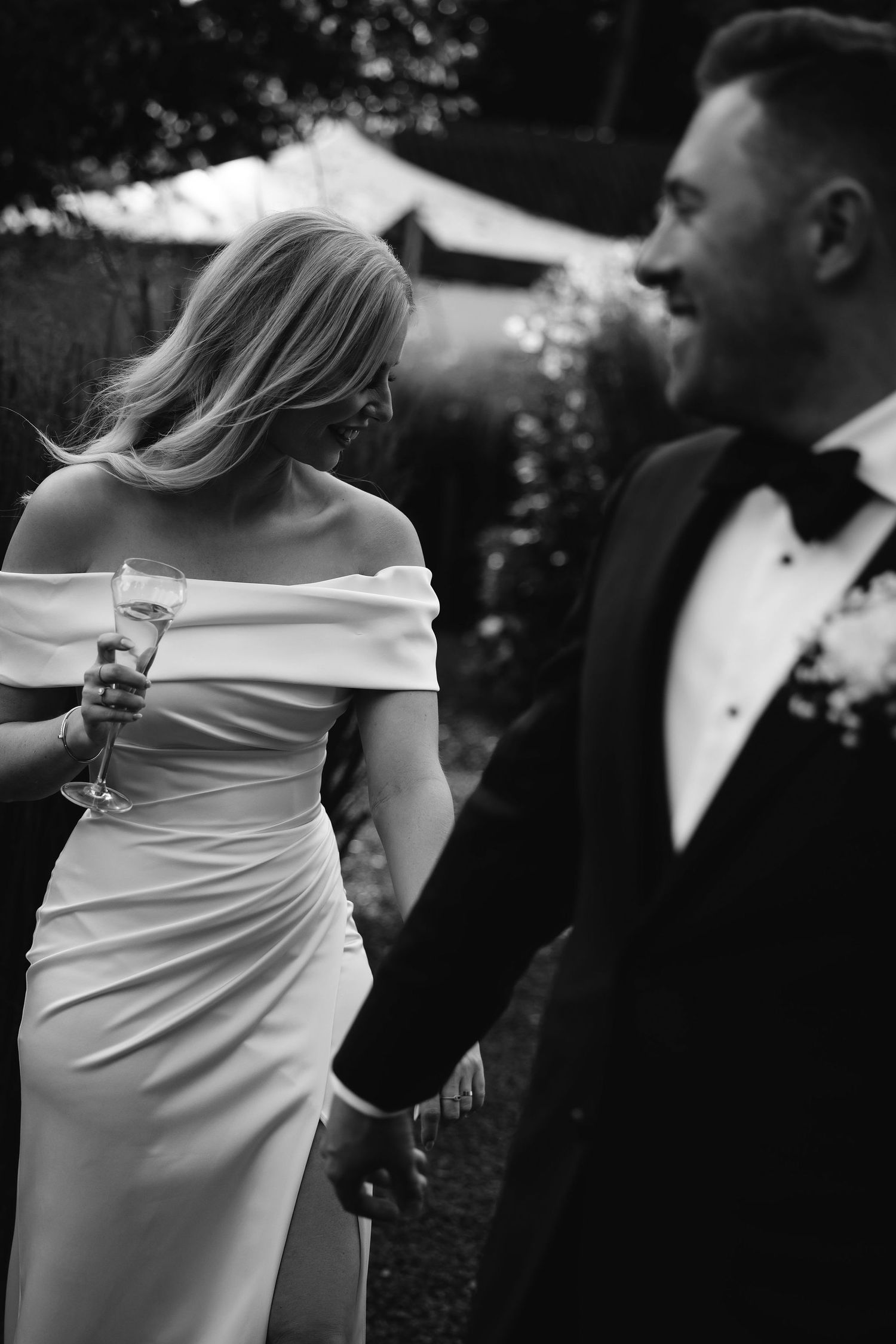 A bride in an elegant off-shoulder white dress holds a champagne glass while celebrating at an outdoor wedding reception.