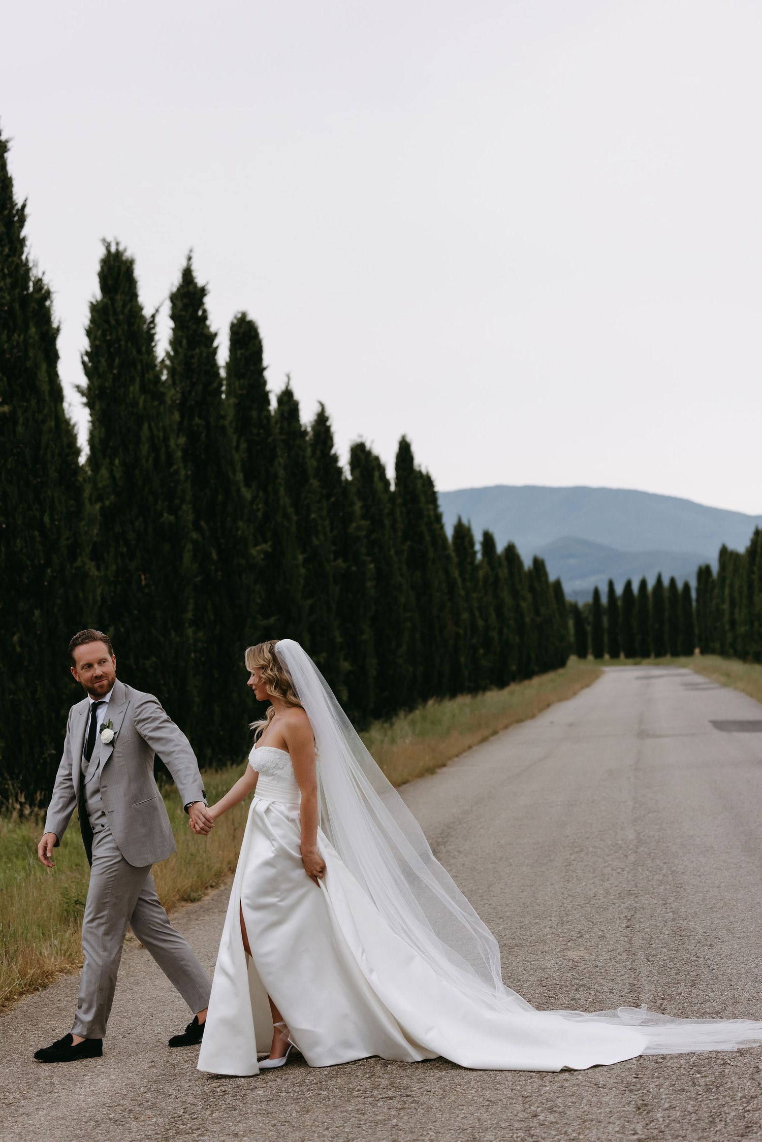 A wedding couple walking down a cypress tree-lined path with mountains in the background.