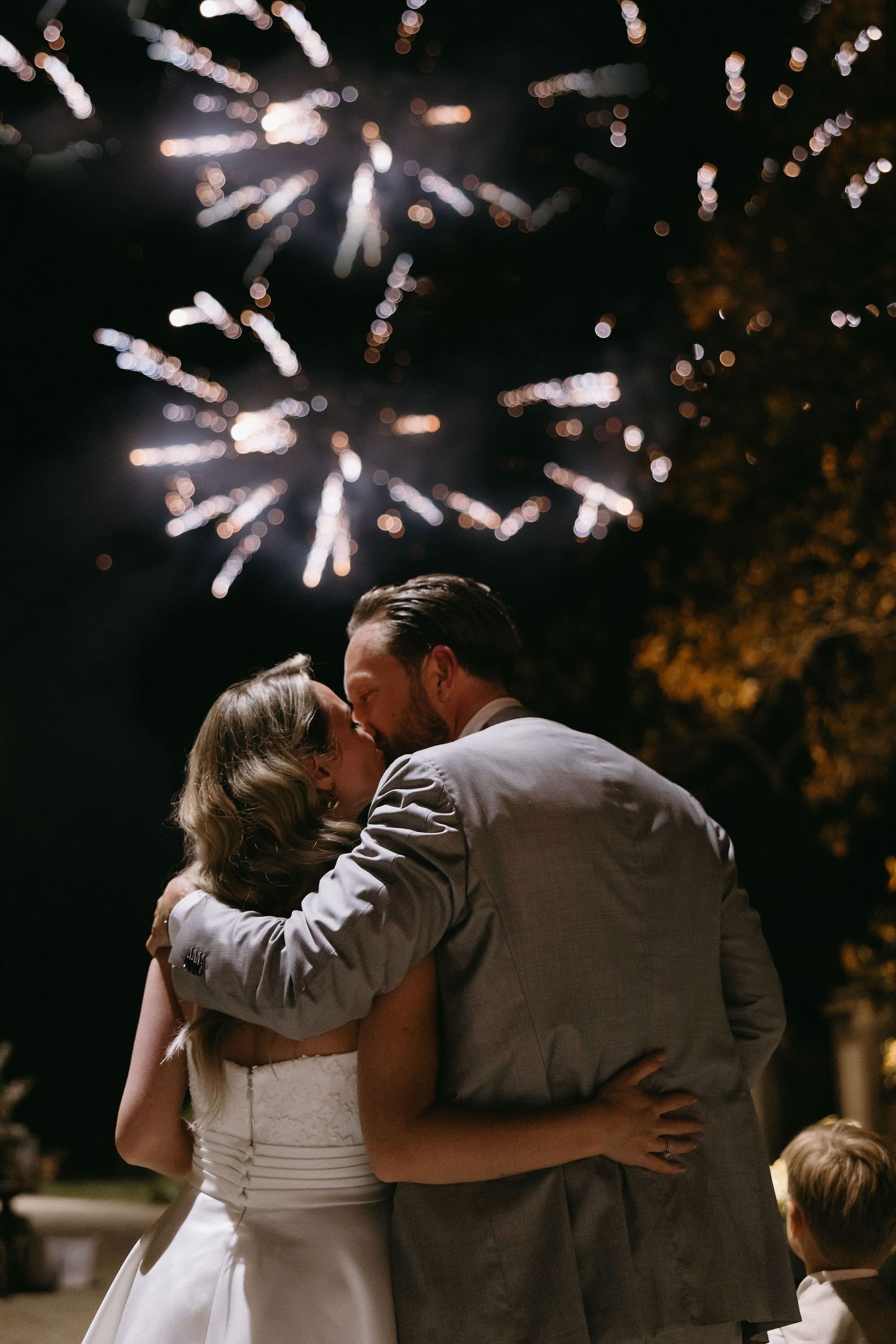 A romantic kiss under bursting fireworks at an outdoor wedding celebration.