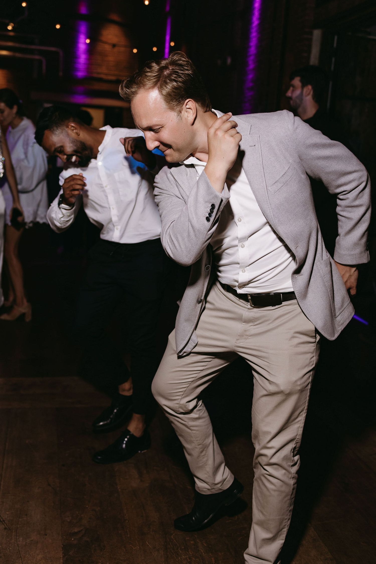 Wedding guests in light colored suits dancing enthusiastically on dark dance floor.
