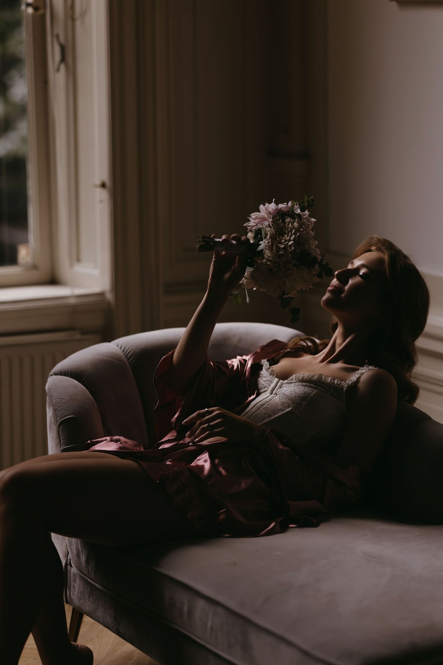 Silhouette reclining on couch holding flowers in dramatic window lighting.