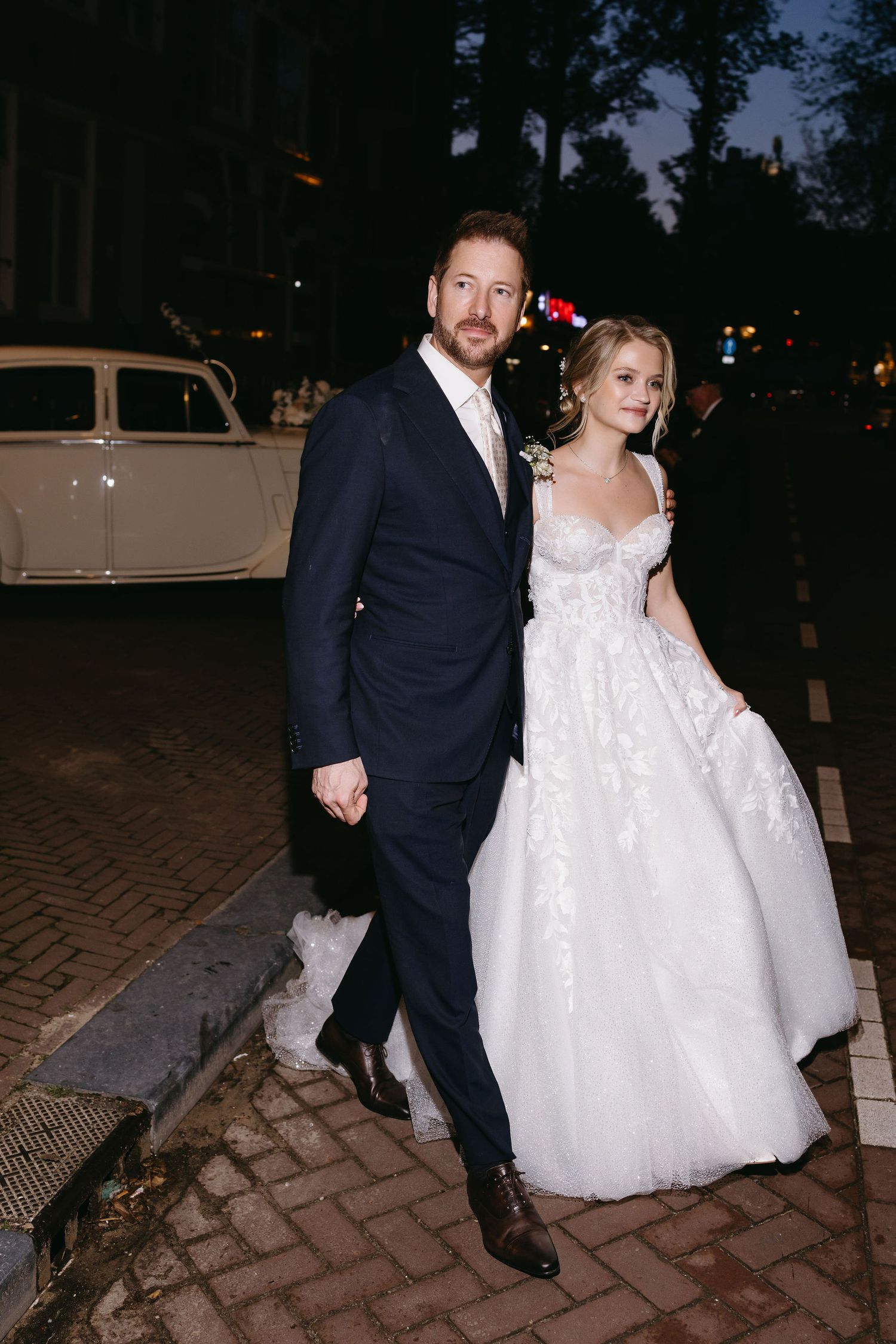 Couple in wedding attire pose together on a cobblestone street at night with a vintage car in background.