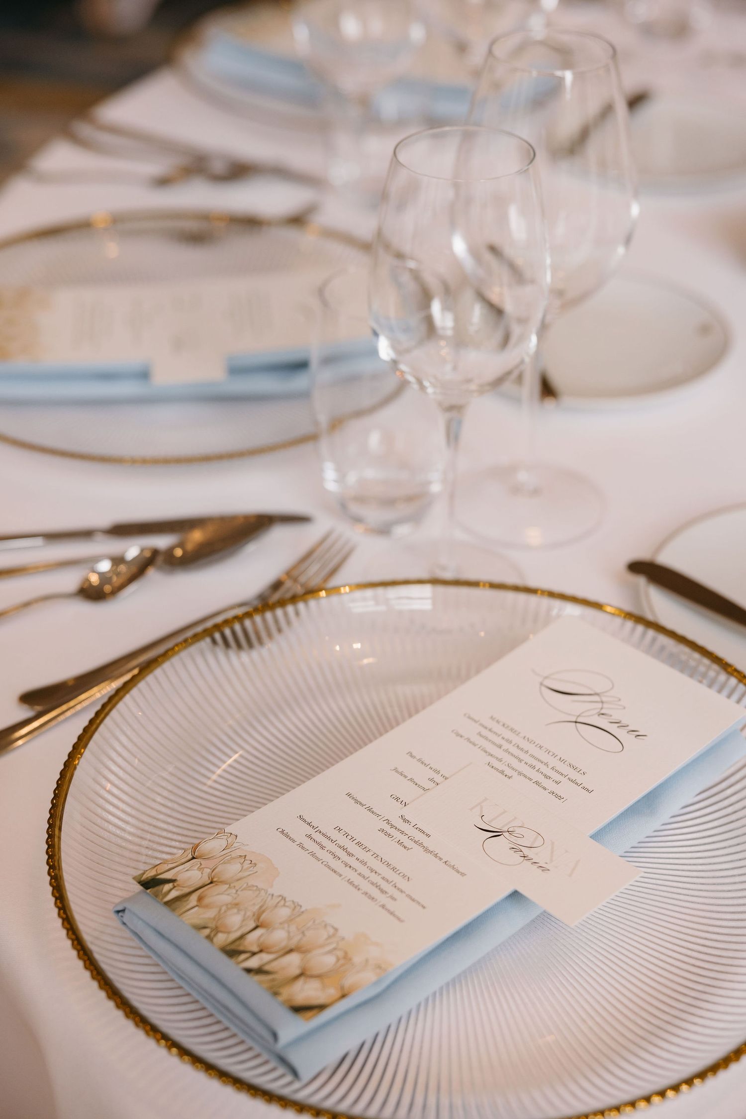 Elegant place setting with gold-rimmed plate, menu card, and stemware on white tablecloth.