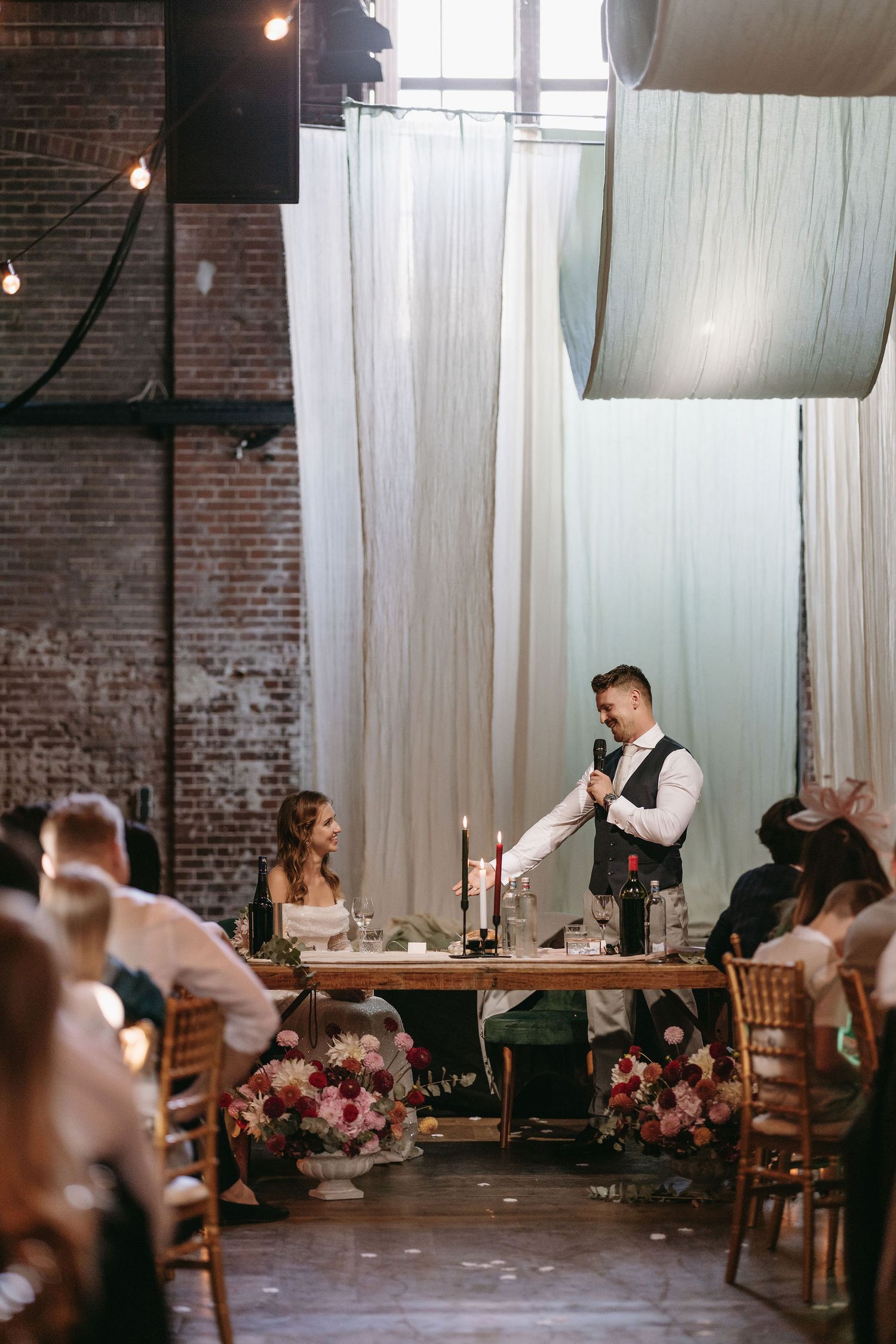 Wedding reception in an industrial brick venue with string lights and white curtain decorations.