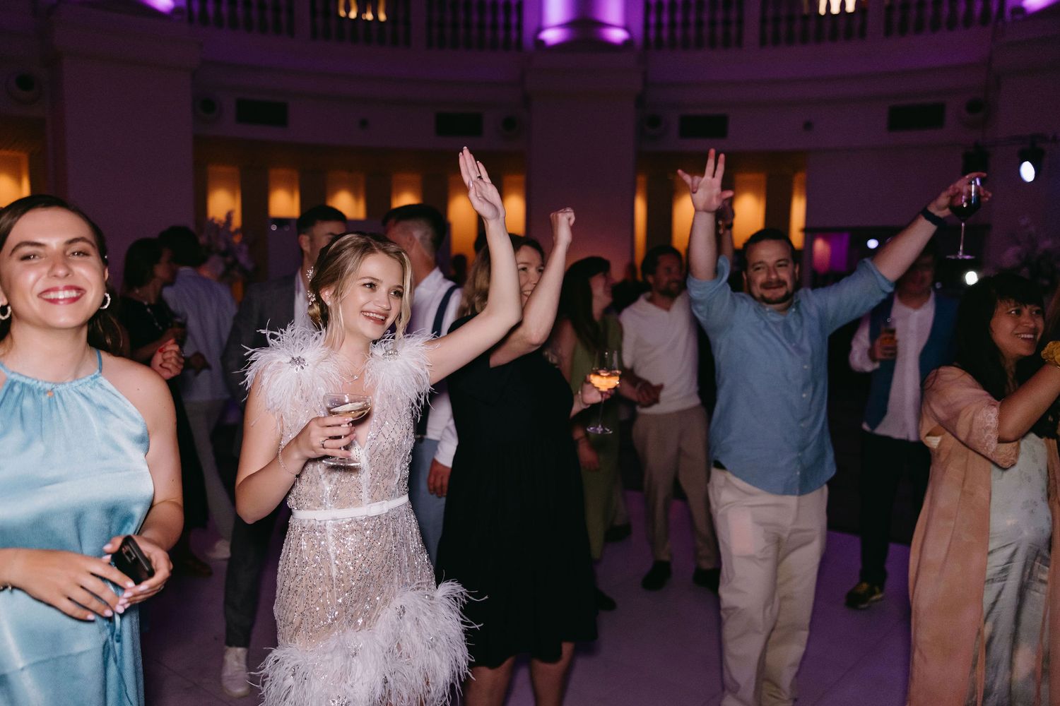 Wedding guests dancing and celebrating at reception with purple lighting.