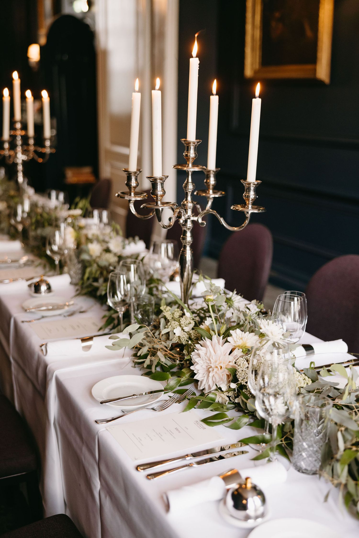 Elegant wedding table setting with white candelabras, floral runner and crystal glassware in a dark moody room.