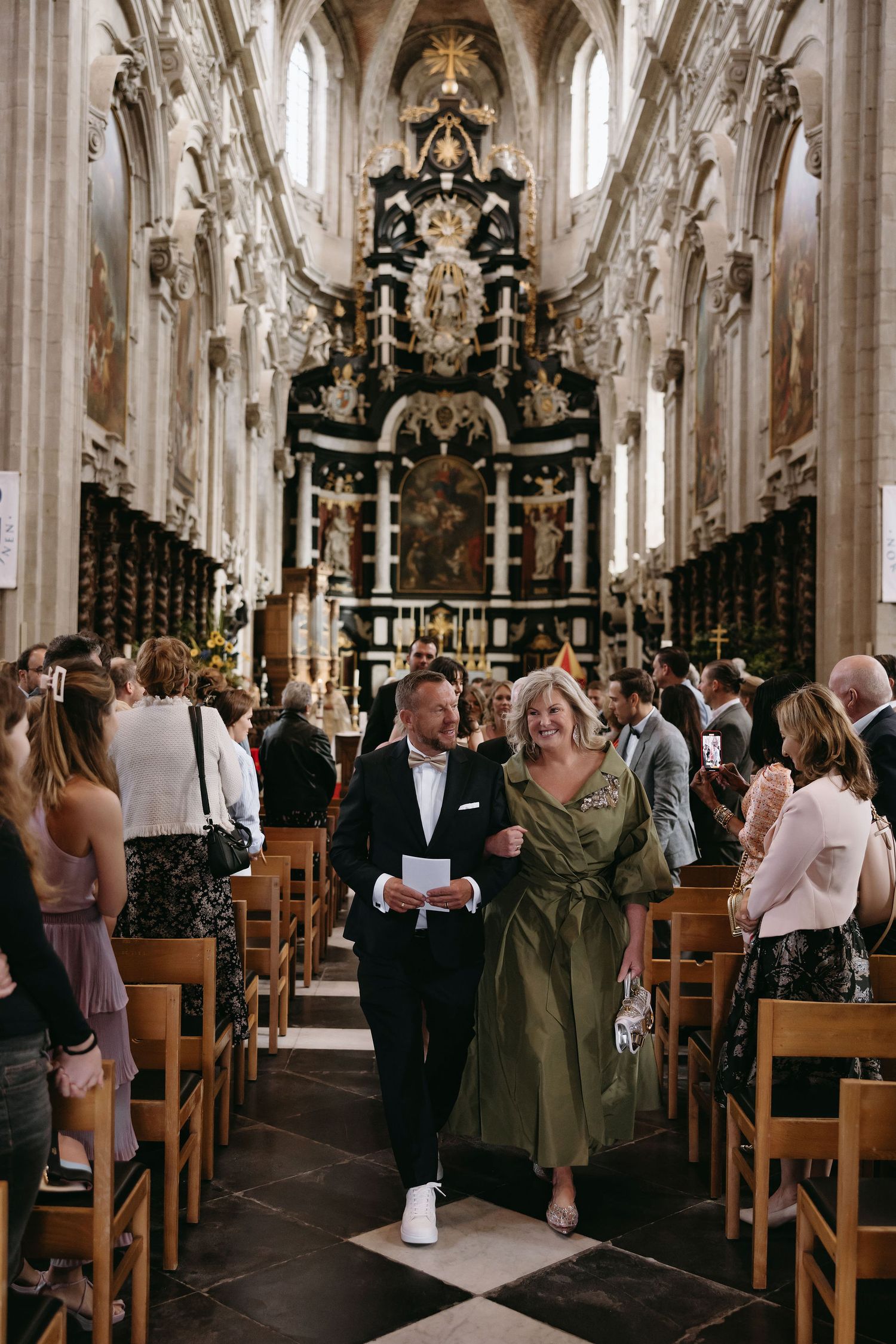 Guests walking down church aisle with ornate baroque altar and architecture.