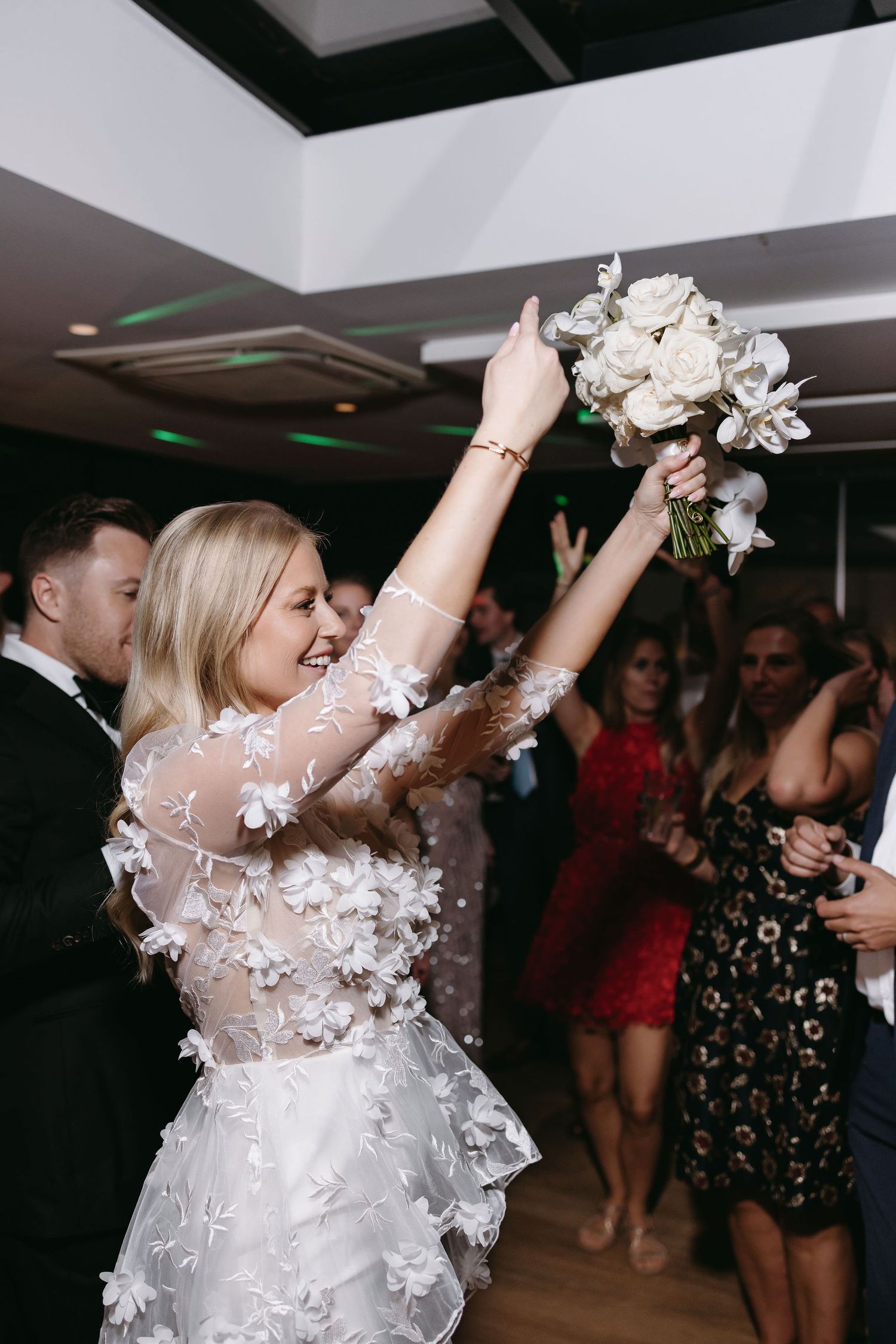 A bride in a floral applique gown raises her bouquet while dancing at a wedding reception.