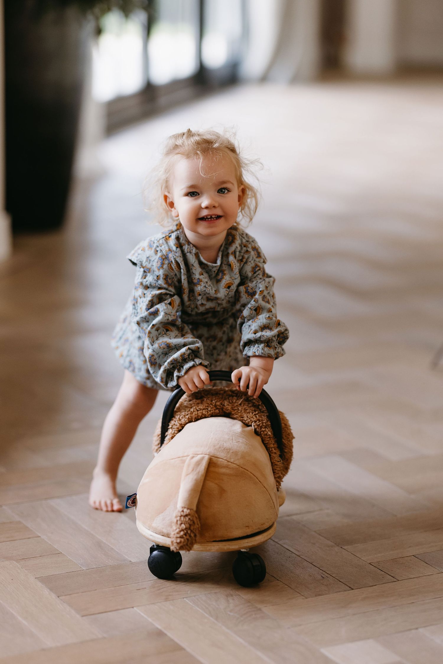 Child in floral dress plays happily with cushion on herringbone floor.