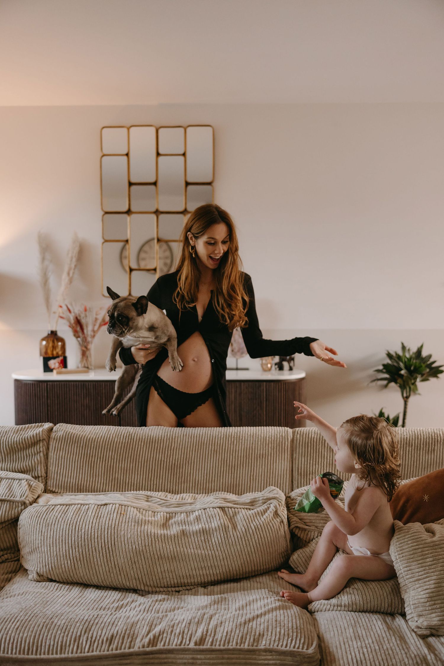 A pregnant woman in black lingerie poses playfully with a dog and toddler on a beige couch.
