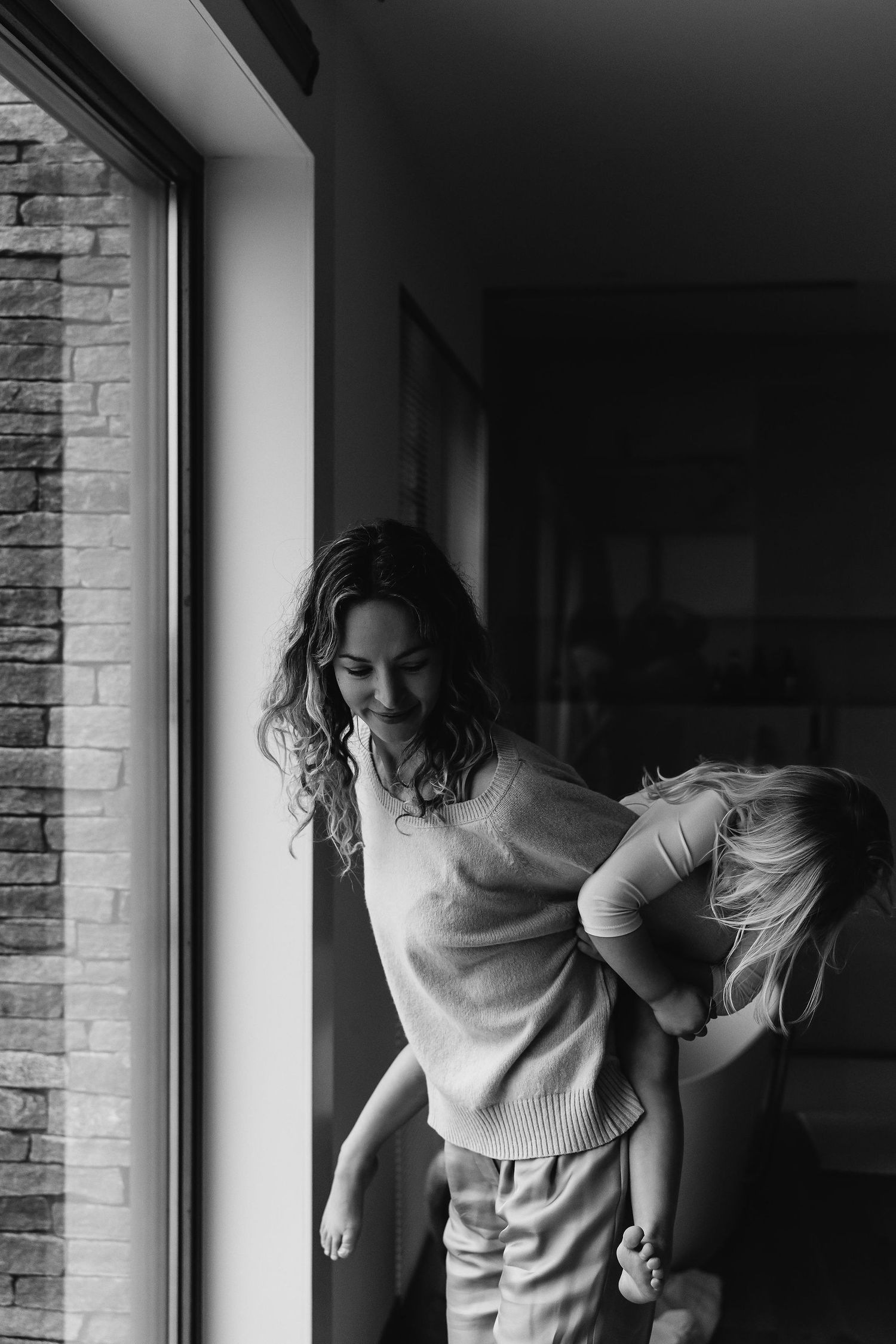 A black and white capture of a playful piggyback ride near a window with brick walls.