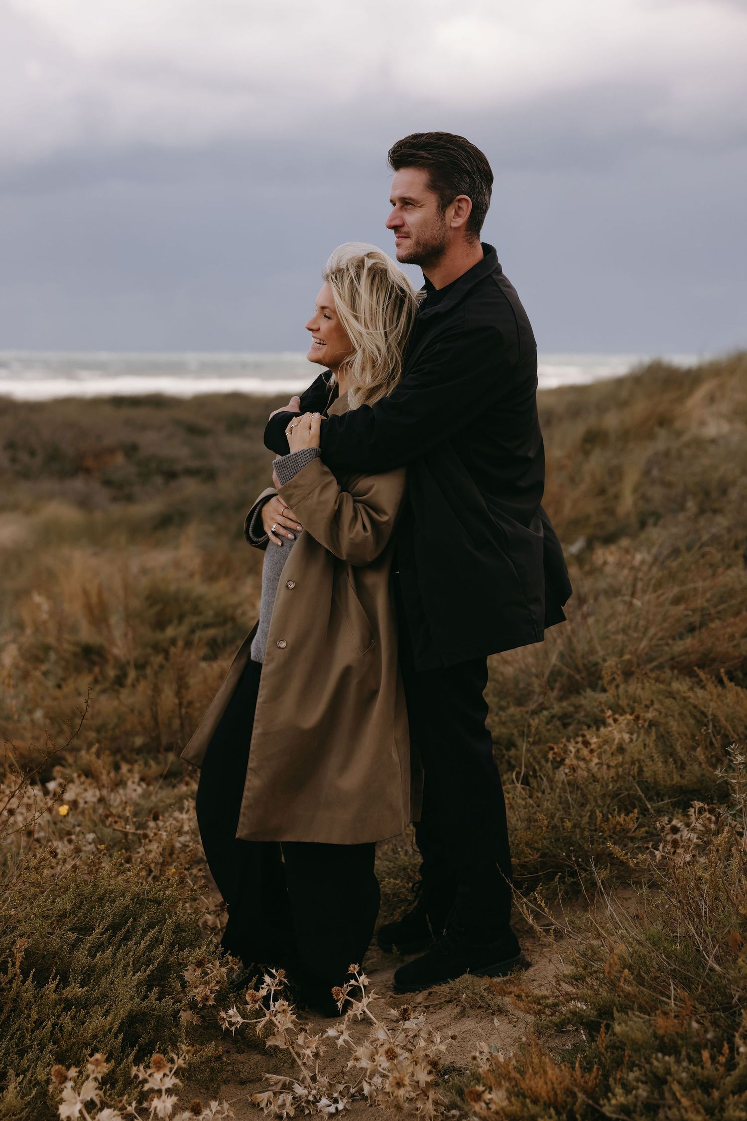 A couple shares an intimate embrace on a coastal cliffside under moody gray skies.