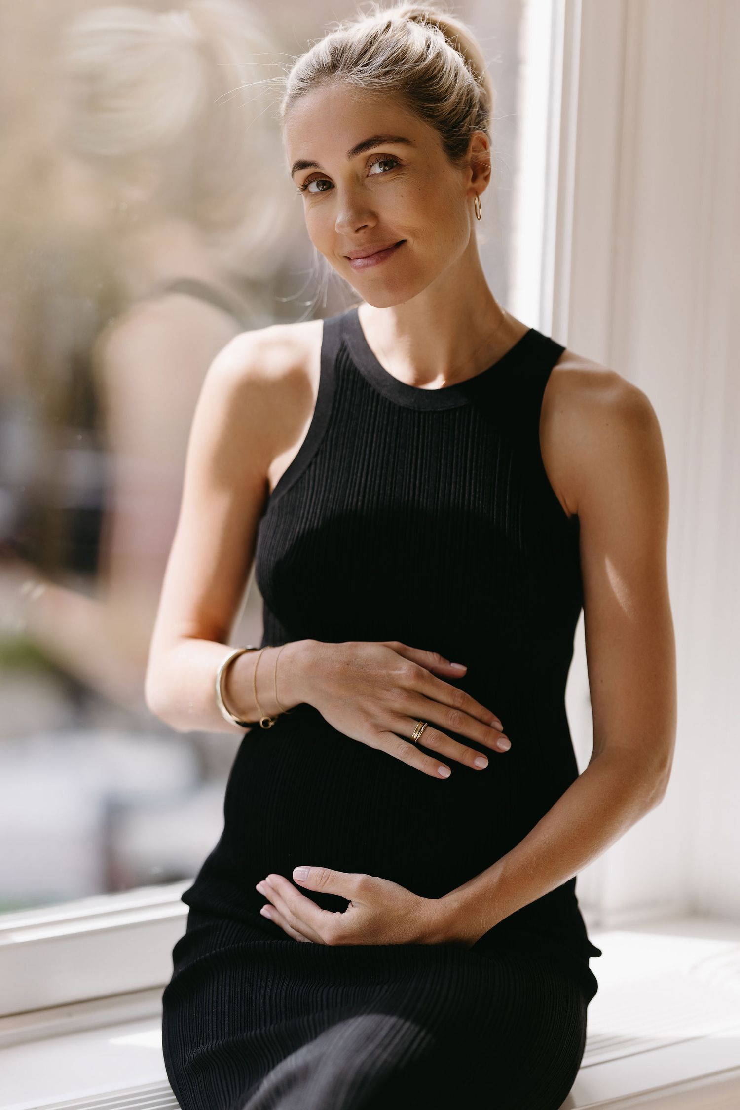 Minimalist maternity portrait in black ribbed tank dress by sunlit window.
