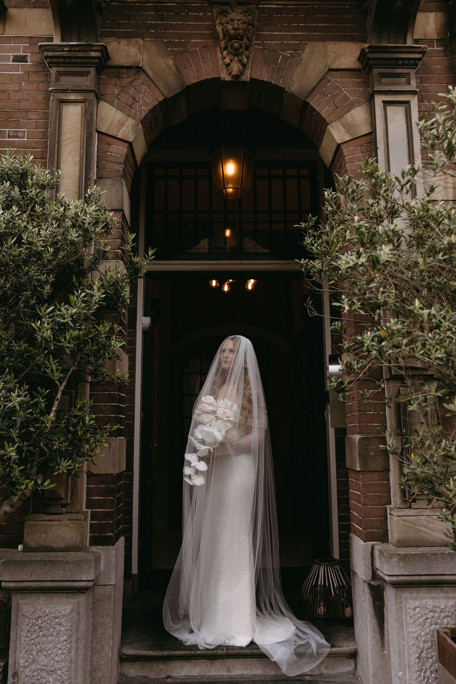 A figure in a long white wedding gown and veil stands elegantly framed in a grand stone archway entrance.