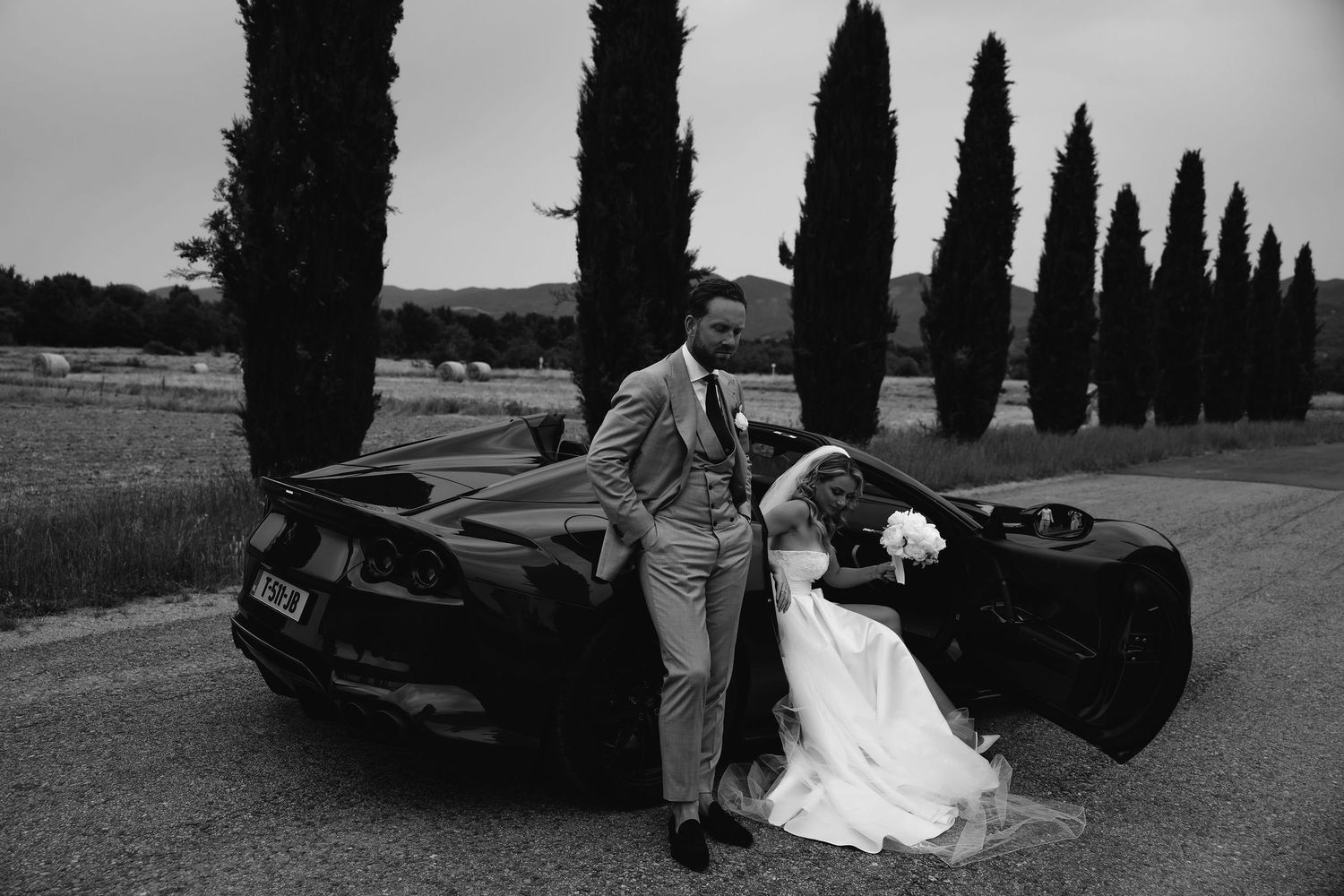 Couple posing by luxury sports car on tree-lined Italian road with tall cypress trees in black and white.