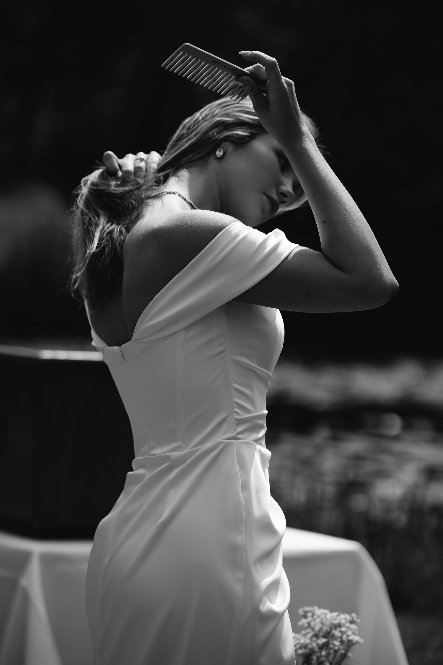 A black and white artistic portrait of a woman in a white dress combing her hair outdoors in dramatic lighting.