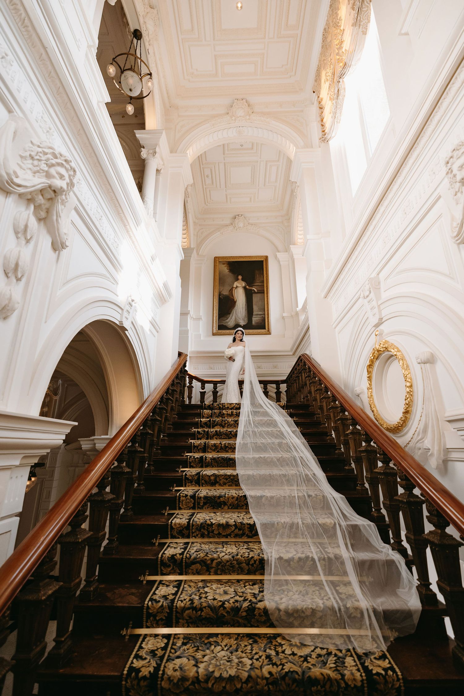 Elegant carpeted staircase in a grand white interior with ornate moldings and a vintage portrait painting at the landing.