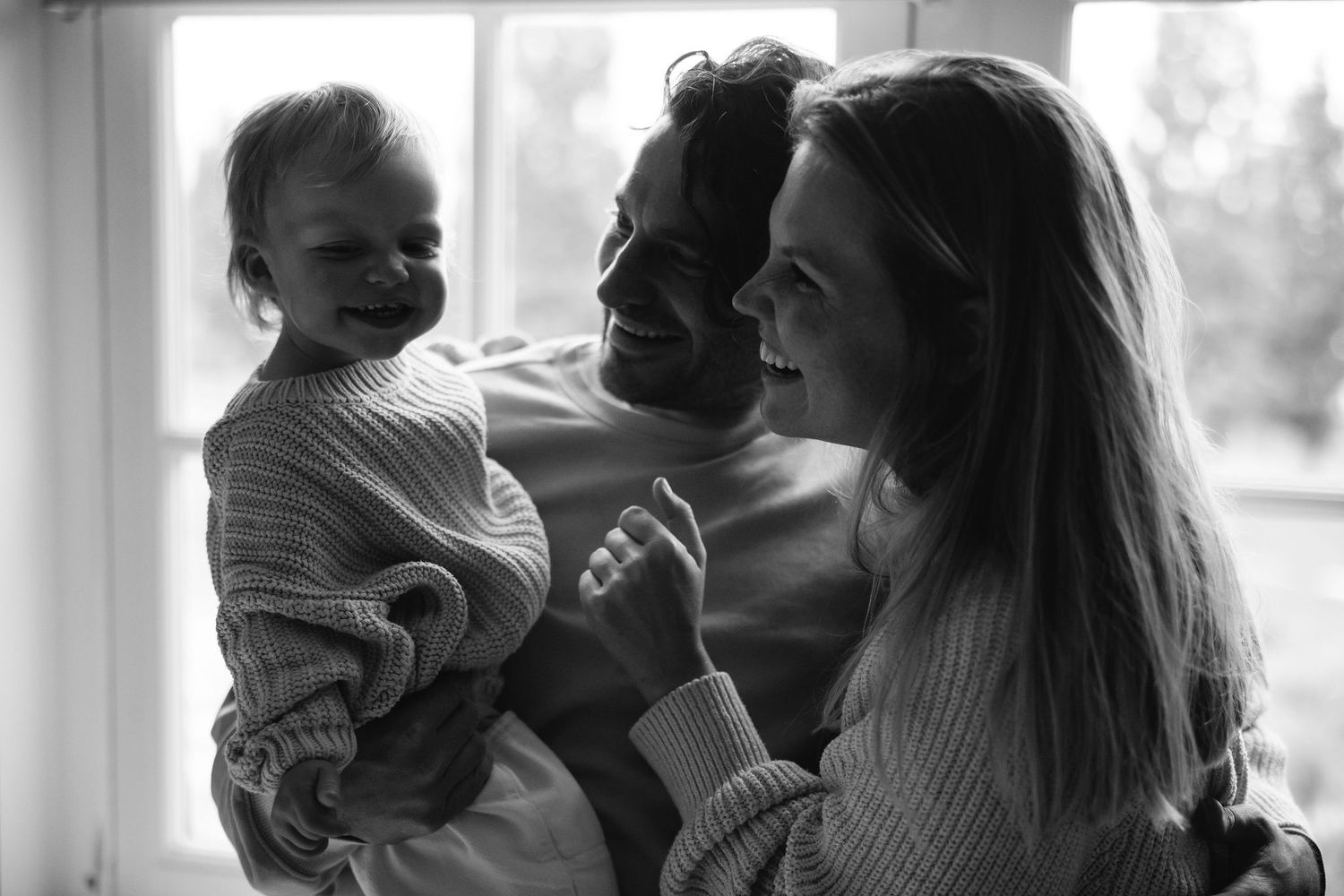 A candid black and white photo of a family laughing together in natural window light.