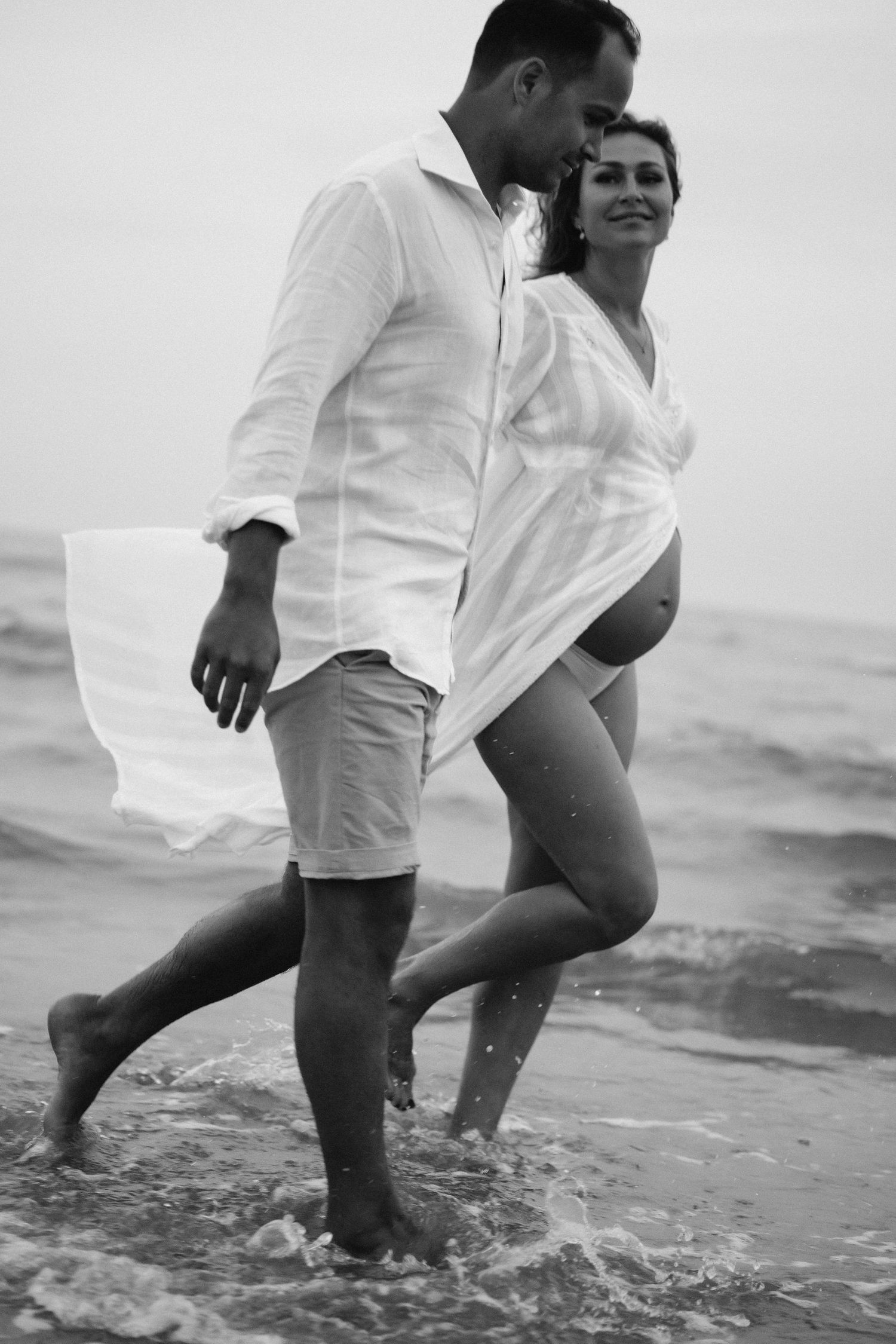A couple in white clothing walks together along the beach, splashing in the shallow waves at the shoreline.