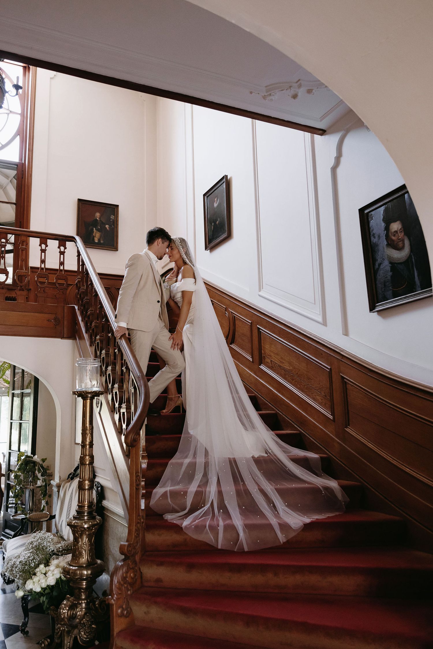 Elegant wedding moment captured on a grand wooden staircase with floral decorations and portrait paintings on the wall.